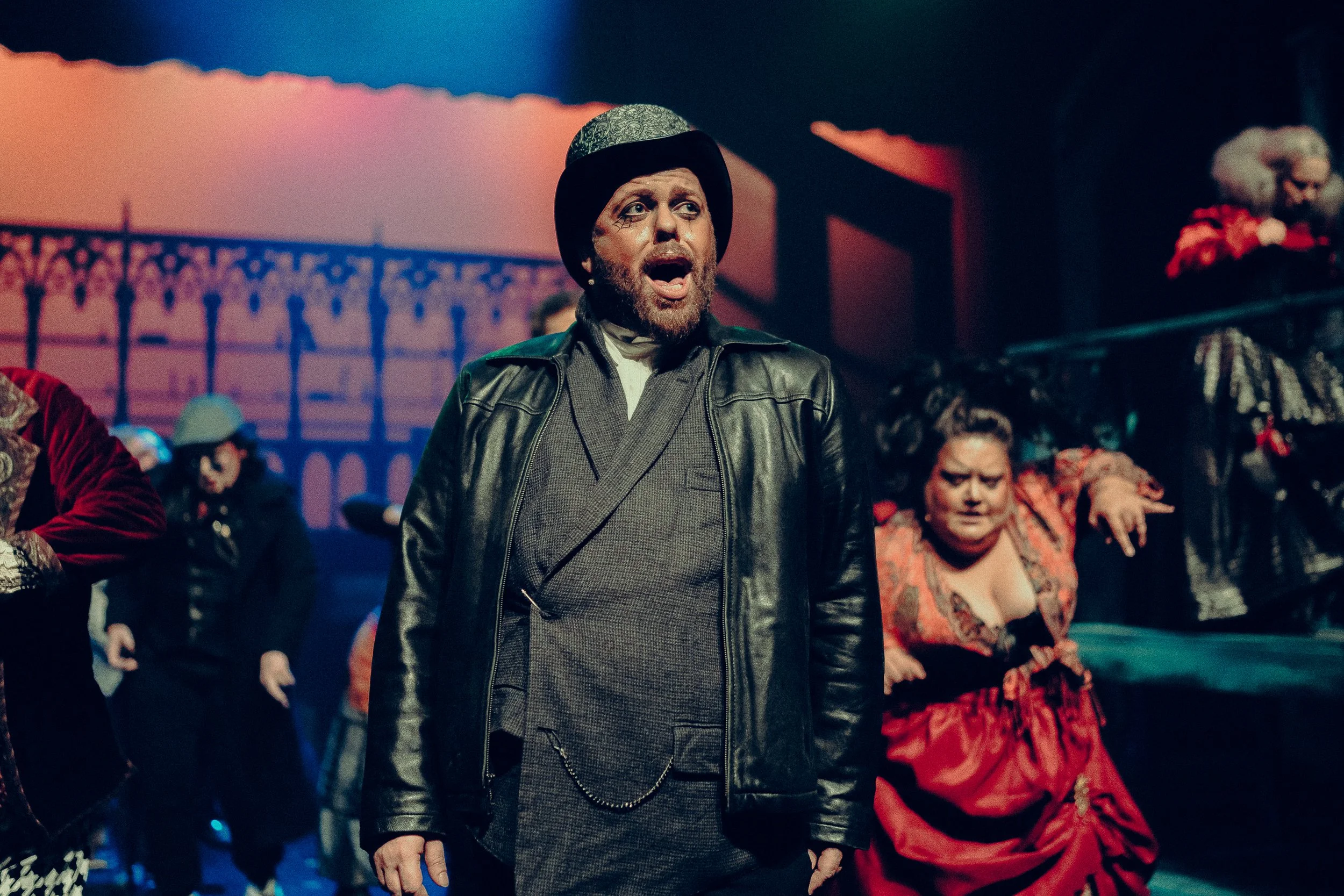 Theatrical performance with actors in period costumes; man in front wearing a leather jacket and bowler hat, others in Victorian-style attire, on stage with dramatic lighting.