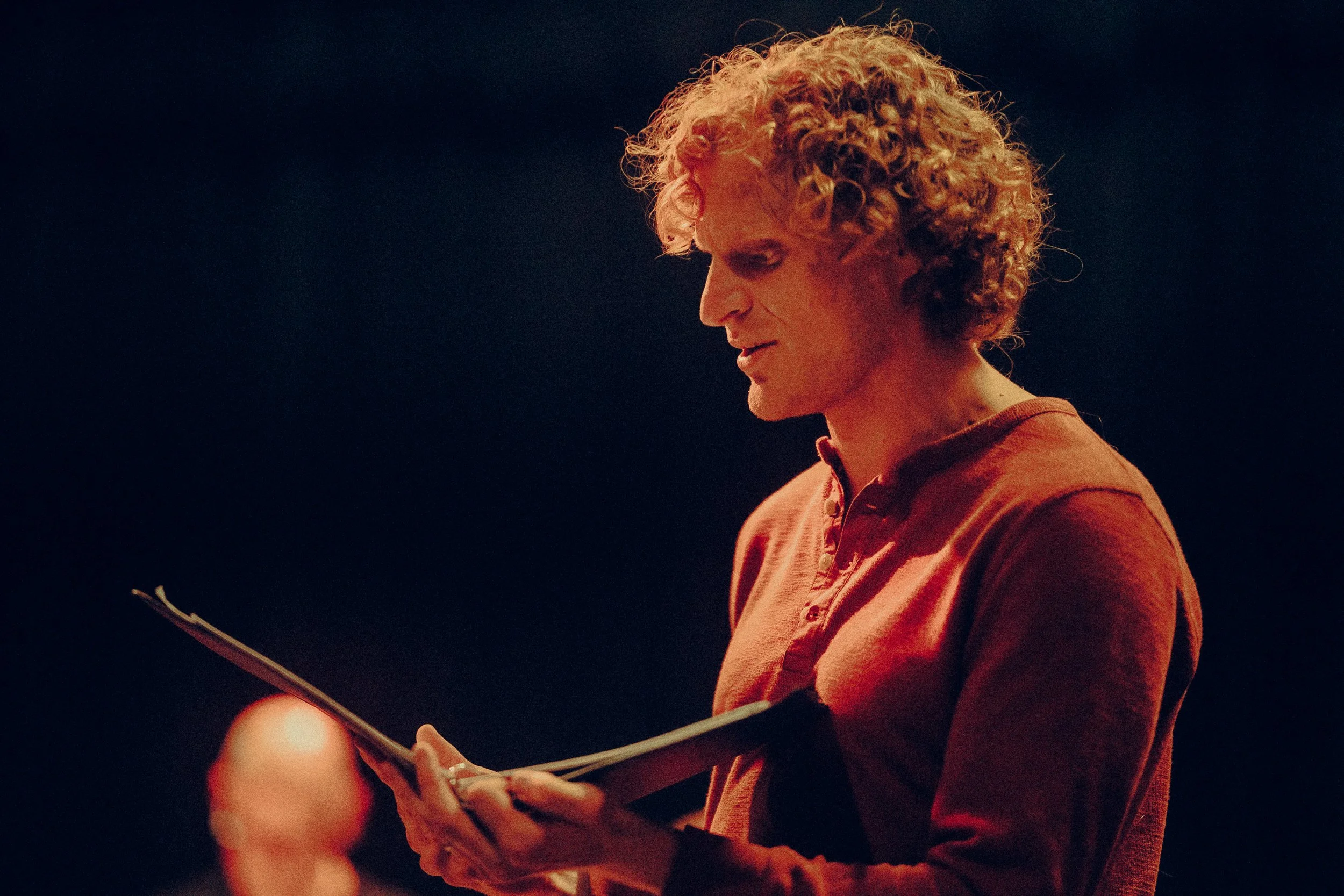 Person in red shirt holding a script, engaged in reading or acting on stage.