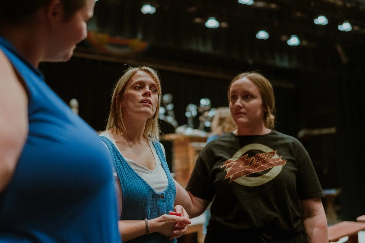 Three women talking in a dimly lit theater or performance space, one wearing a blue top, another in a black shirt, and one holding a red object.