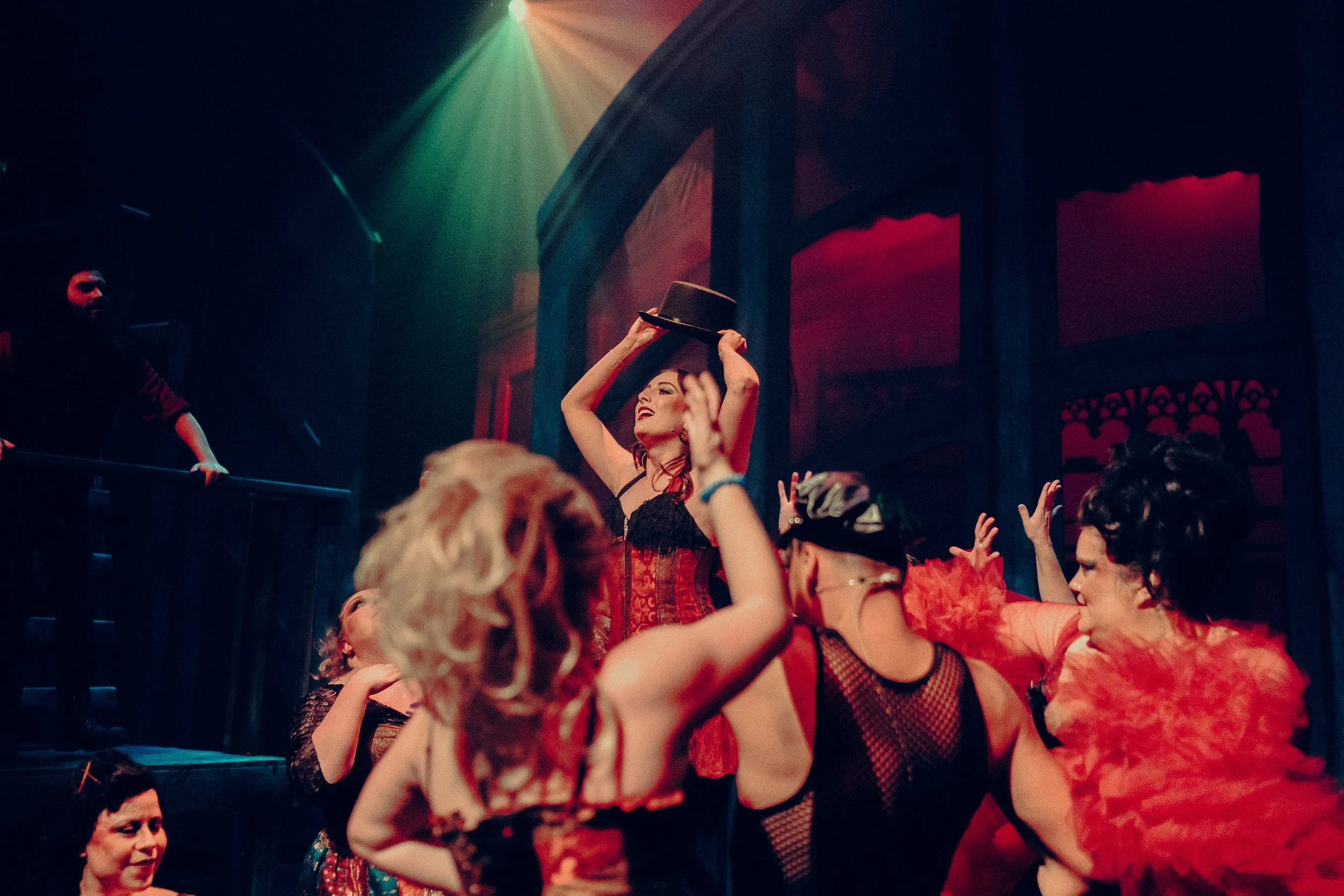 Stage performance with people in theatrical costumes, including corsets and feathered accessories, under red and orange lighting.