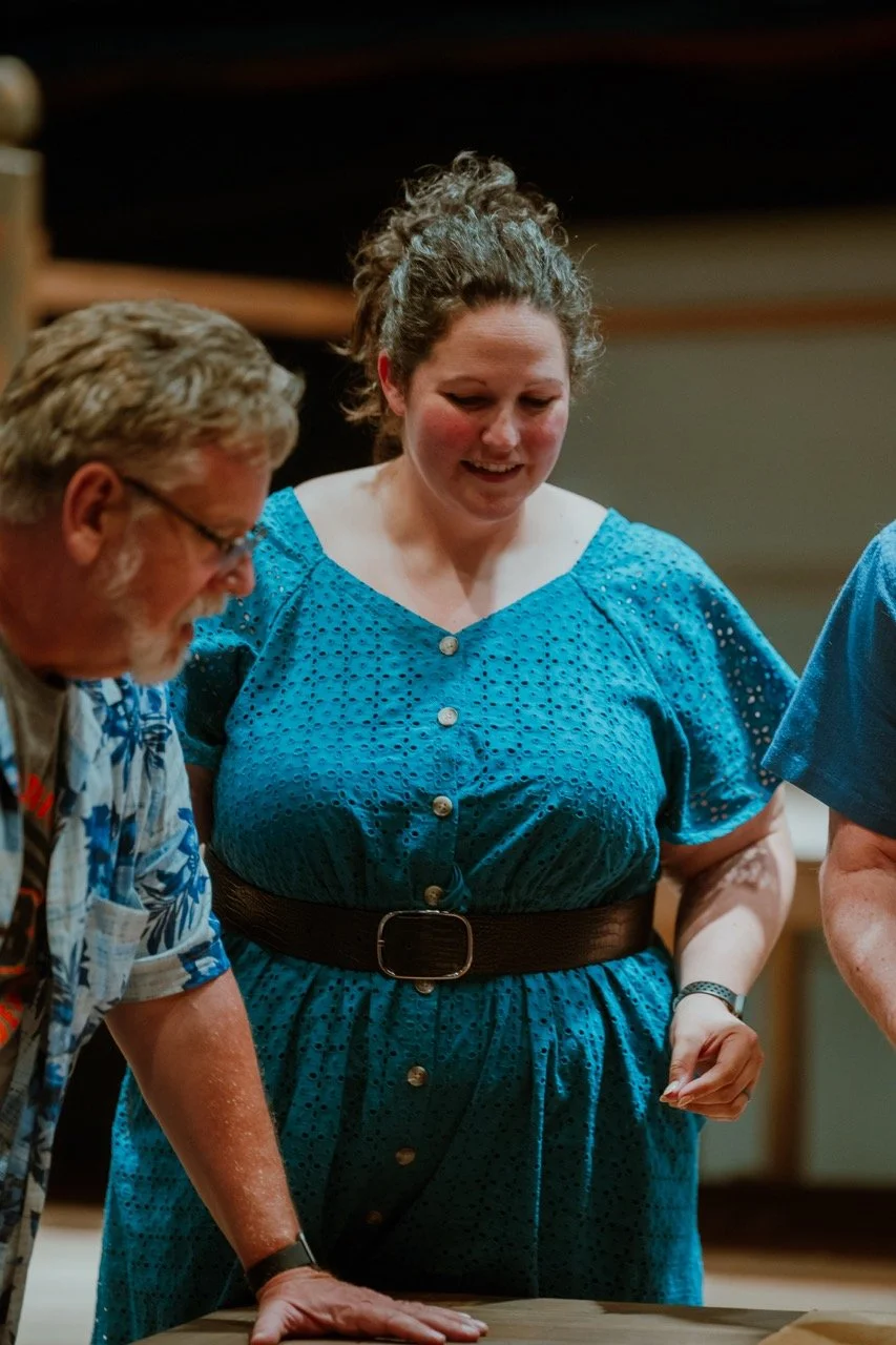 A woman in a blue dress with a brown belt, smiling and looking down, standing next to a man wearing glasses and a floral shirt.