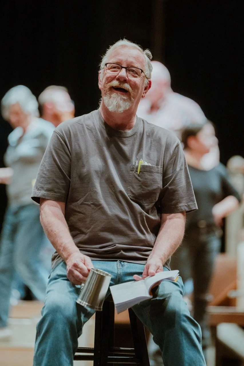 A seated man with glasses and a gray beard is smiling, holding a metal cup and an open book. He is wearing jeans and a gray t-shirt with a pen in the pocket. Blurred people are seen in the background.