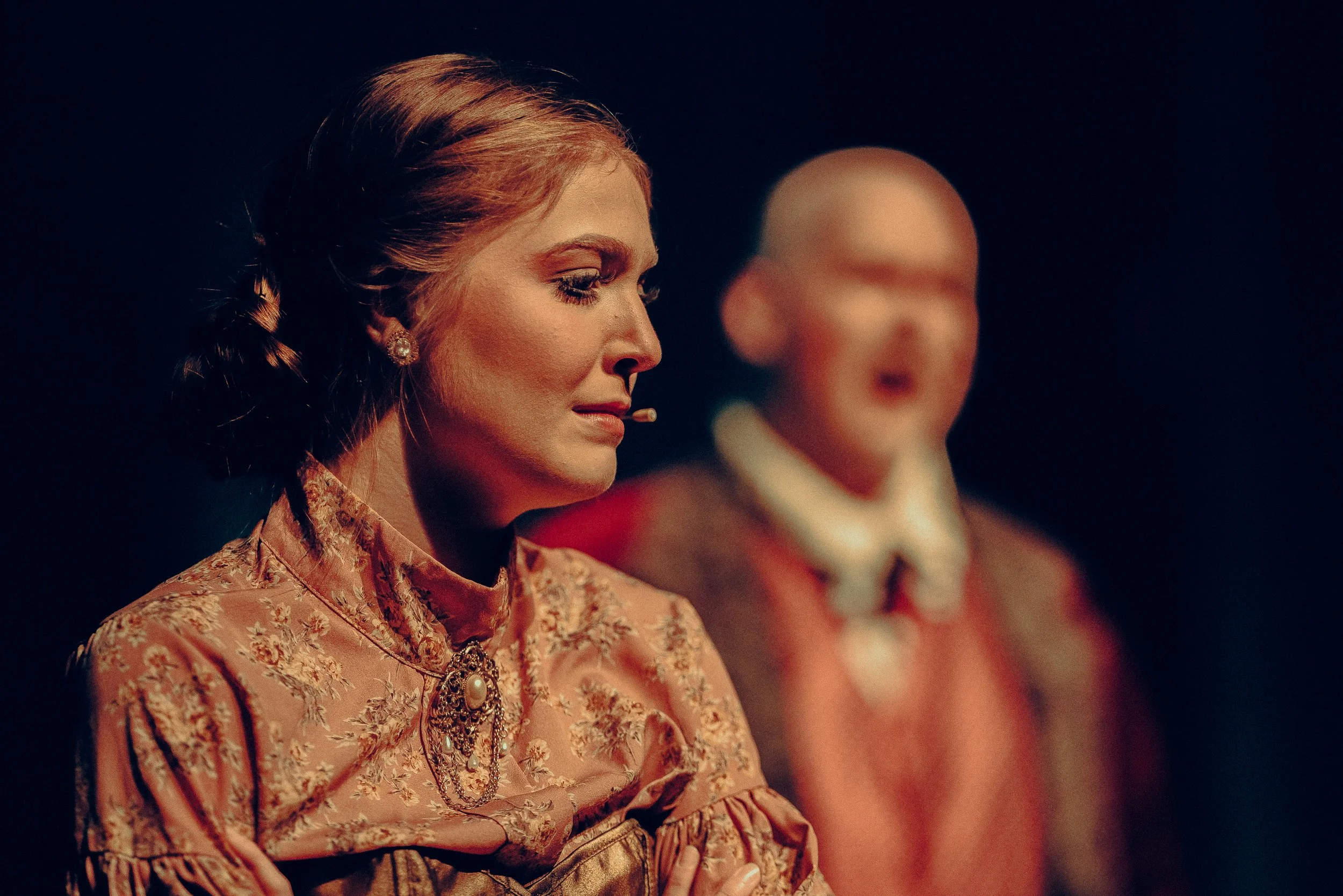A woman in vintage costume with a microphone, focused, with a blurred person in the background on stage.