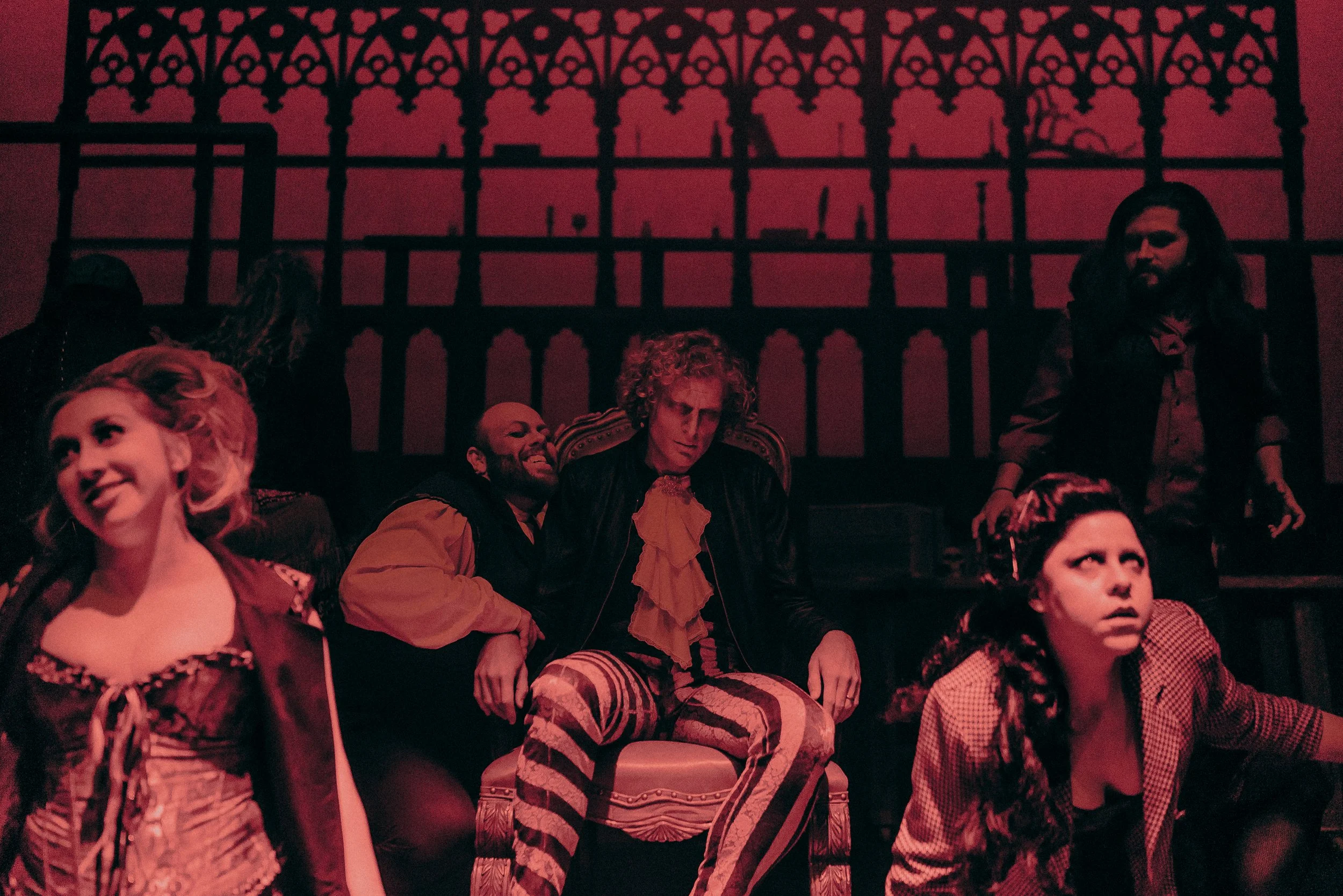 Theatrical performance with actors in dramatic costumes and red lighting. Central figure in colorful pants on a chair, others exhibiting expressive emotions.