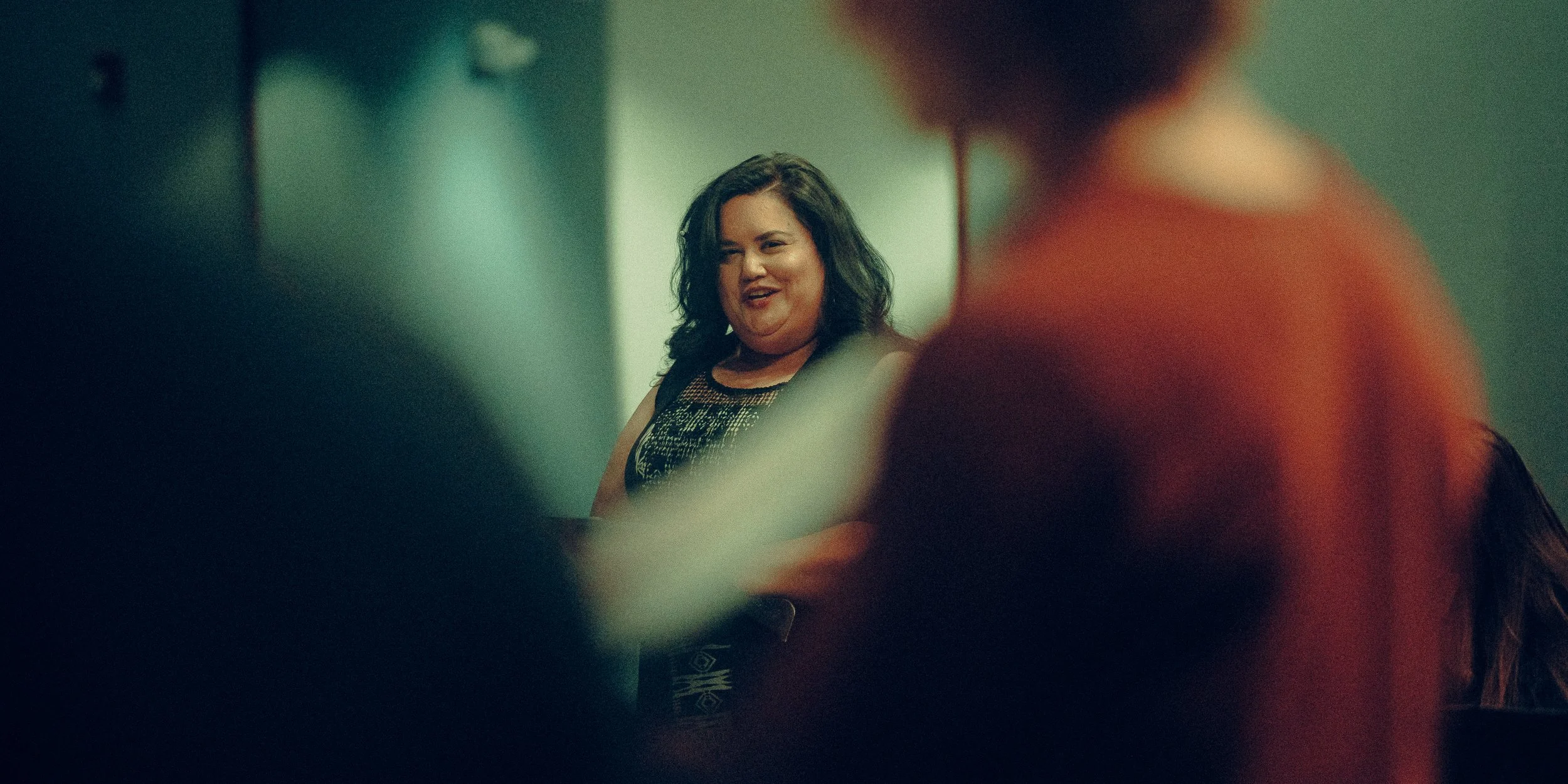 A person smiling, standing indoors with blurred figures in the foreground, creating a depth of field effect.