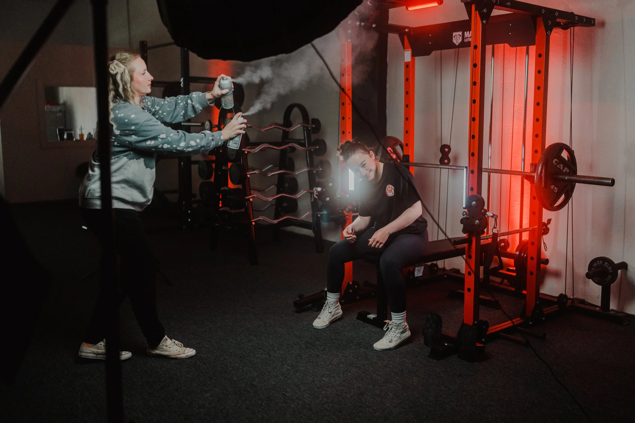 Two people in a gym setting; one is seated on a bench next to weights, while the other sprays mist from a canister.