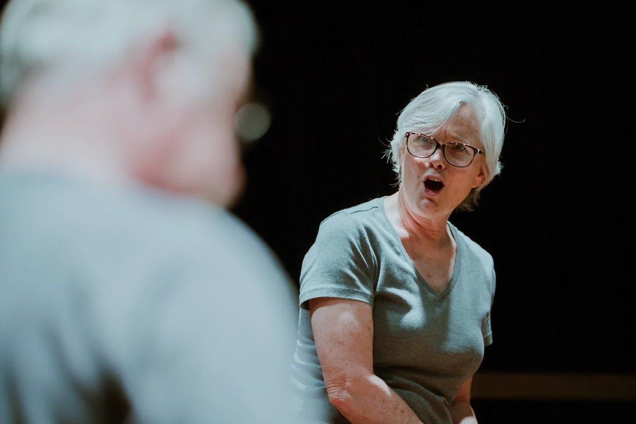 Elderly woman in gray shirt, surprised expression on face, blurred background.