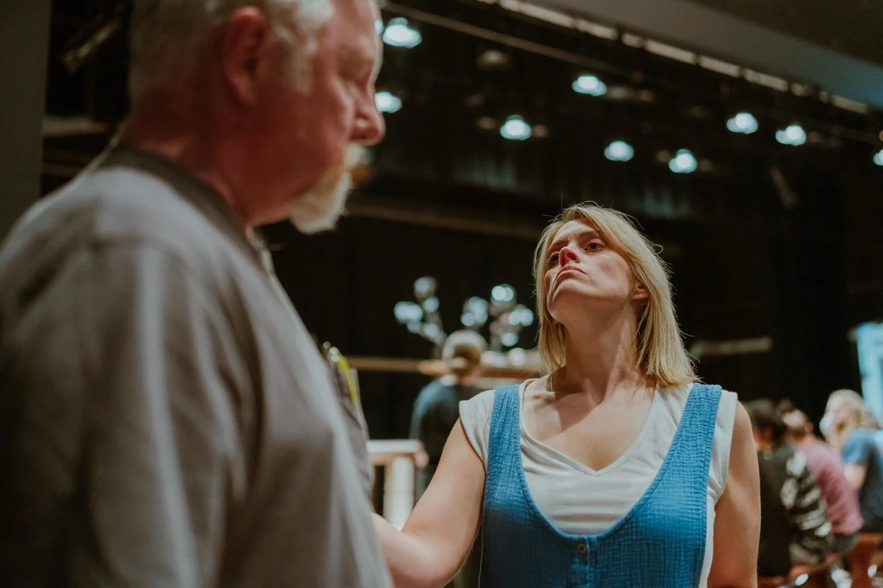 A theater scene with a man and woman in conversation on stage, under bright lights, with other people in the background.