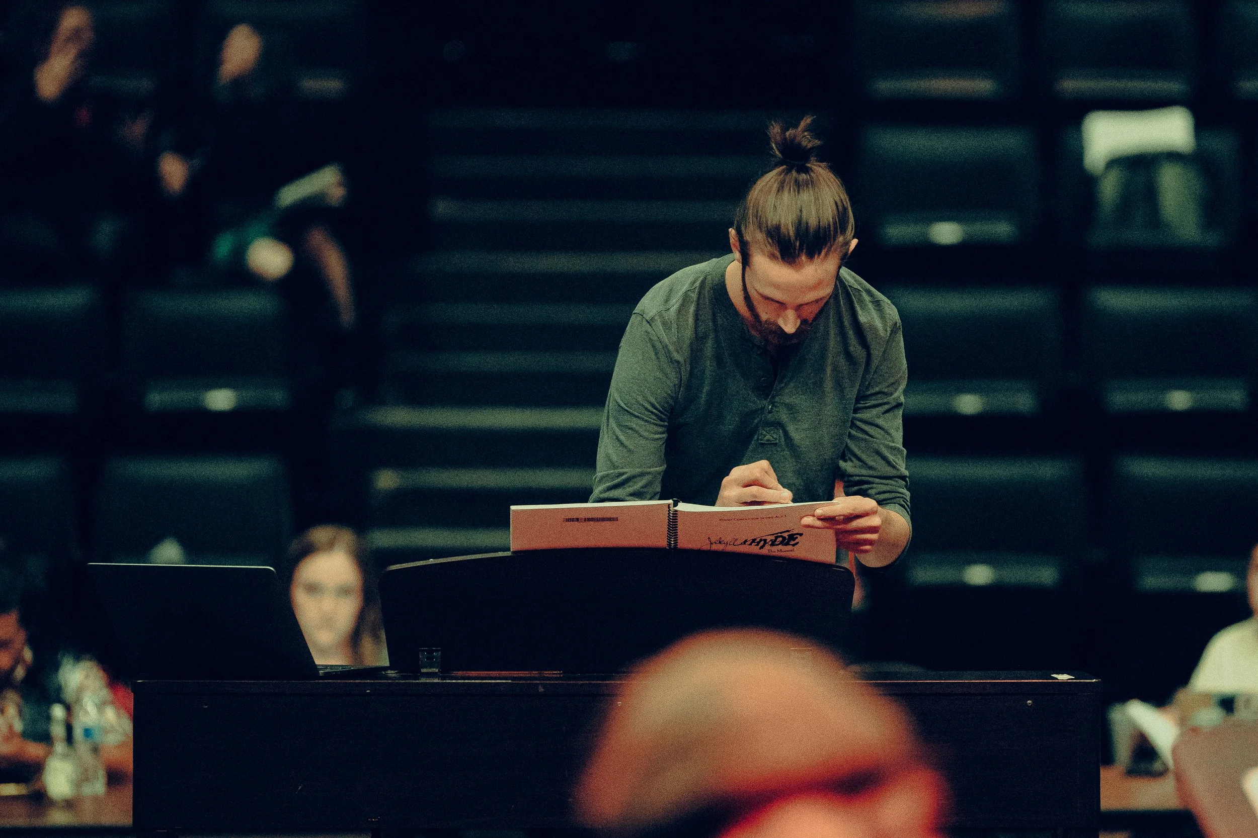 A person with a man bun writing in a notebook at a desk, with a blurred auditorium background.