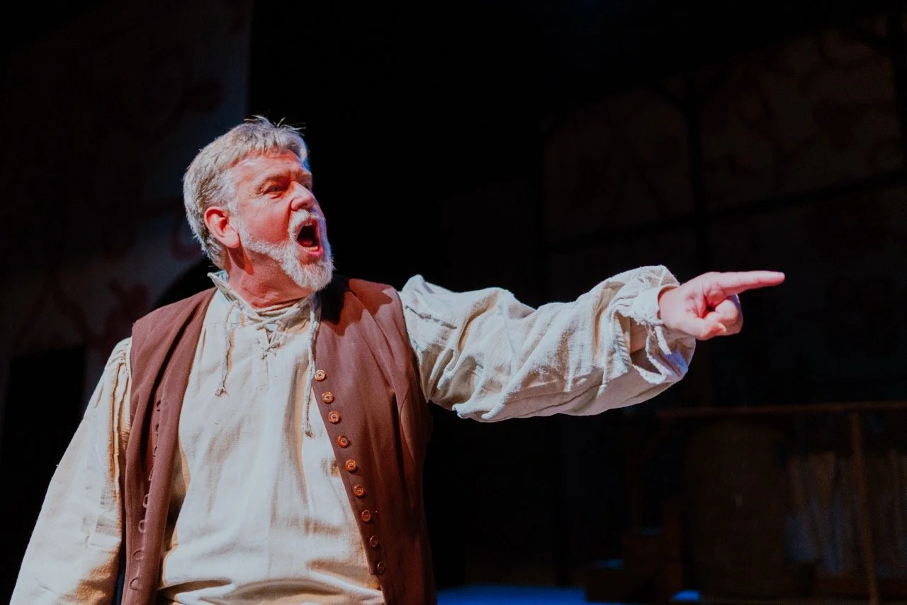 A man performing on stage pointing with an intense expression, wearing a historical costume with a vest and shirt.