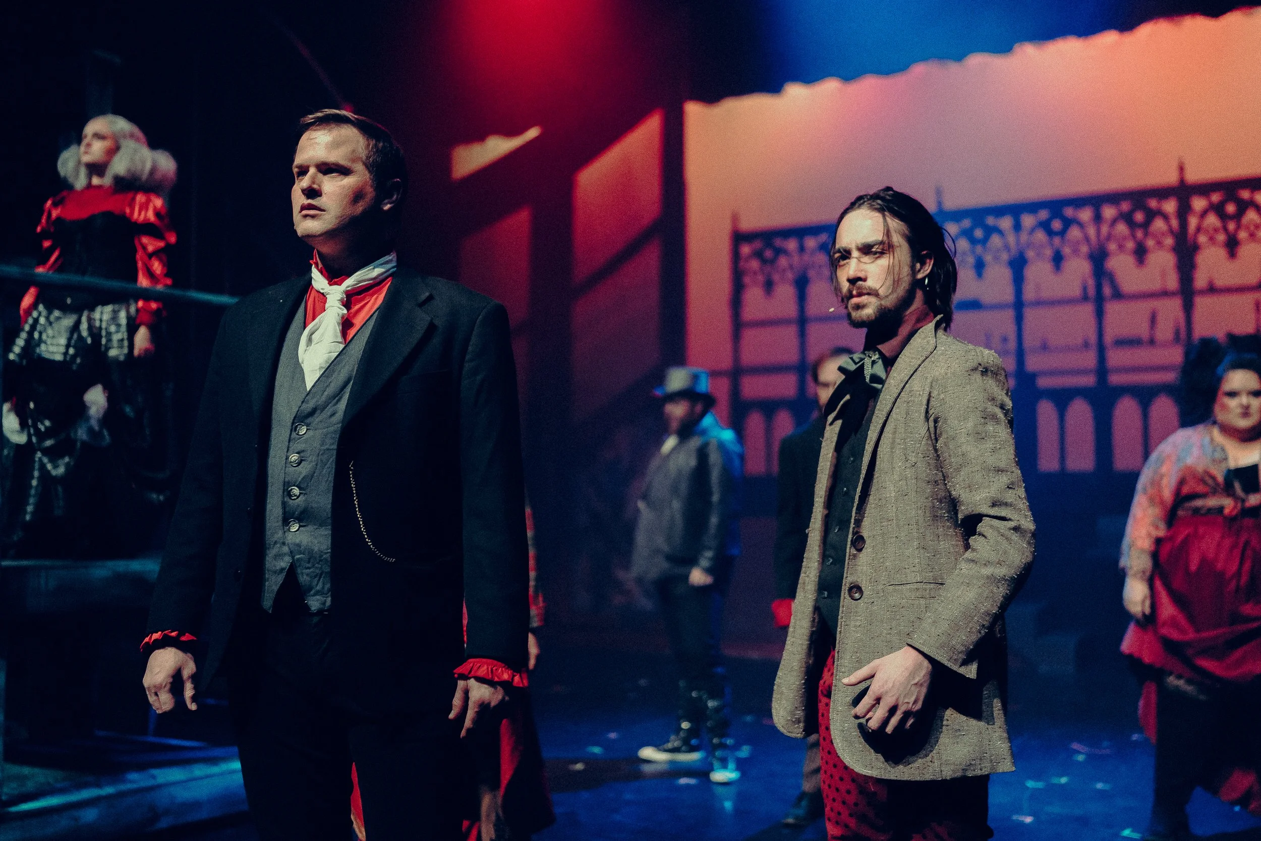 Stage performance scene with actors in Victorian-style costumes, featuring dramatic lighting and a gothic backdrop. Main focus on two men in period attire.