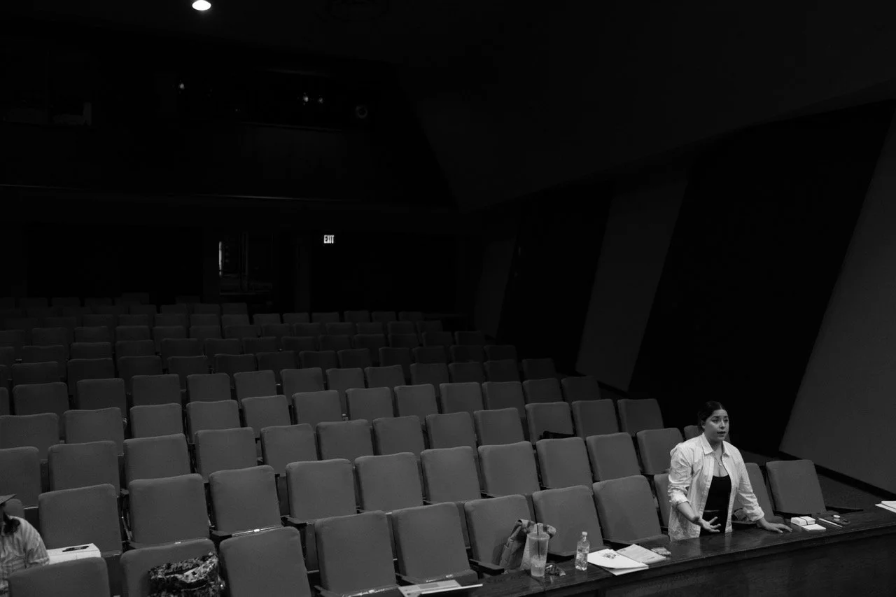 A black and white image of a mostly empty auditorium. A person stands near the front, talking or addressing an audience. Empty seats fill the room, with some items placed on a desk in the foreground.