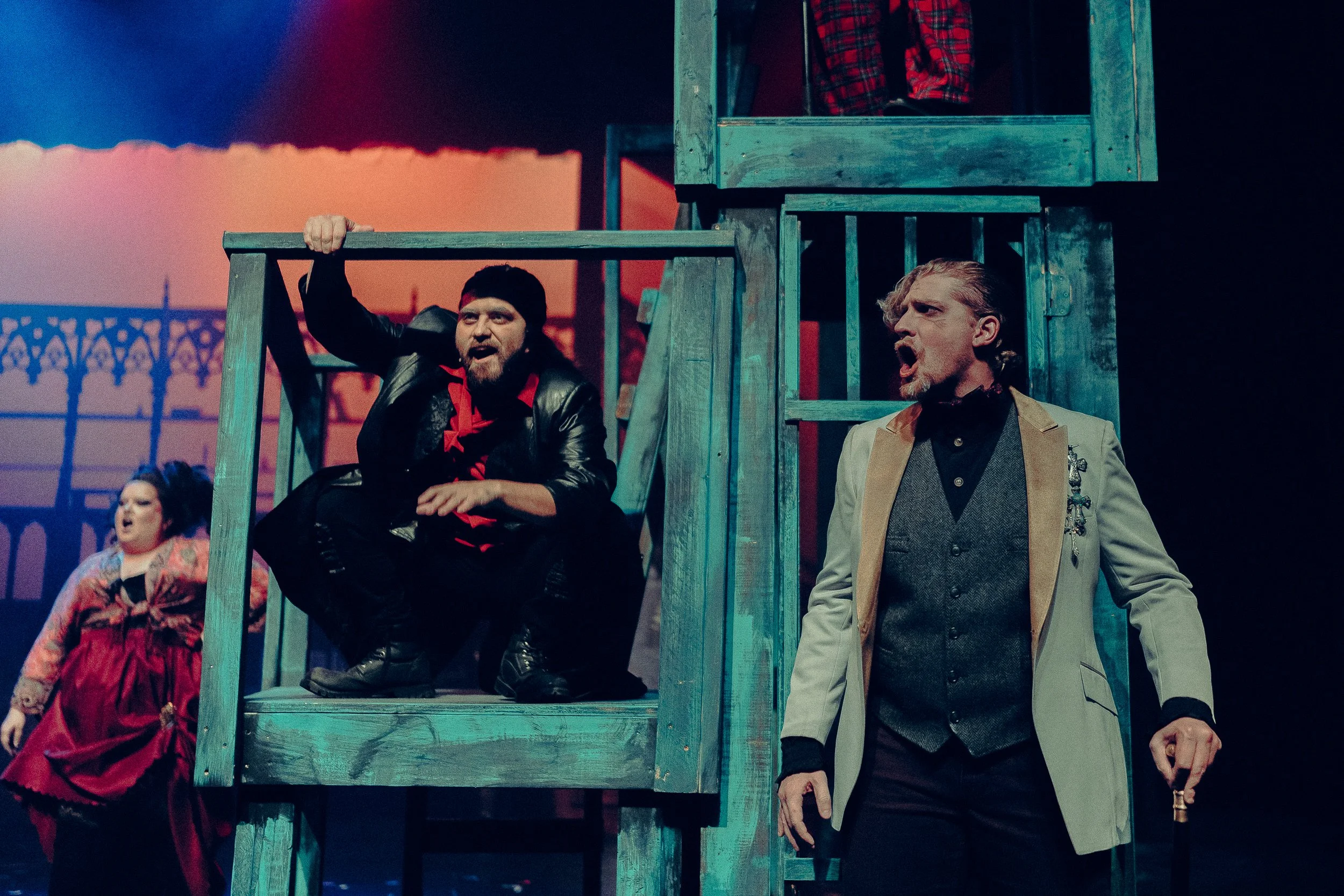 Actors performing on stage in a theatrical play, with one actor squatting on a blue structure and another standing and singing. A woman in a red dress is in the background, and the stage is lit with colorful lighting.