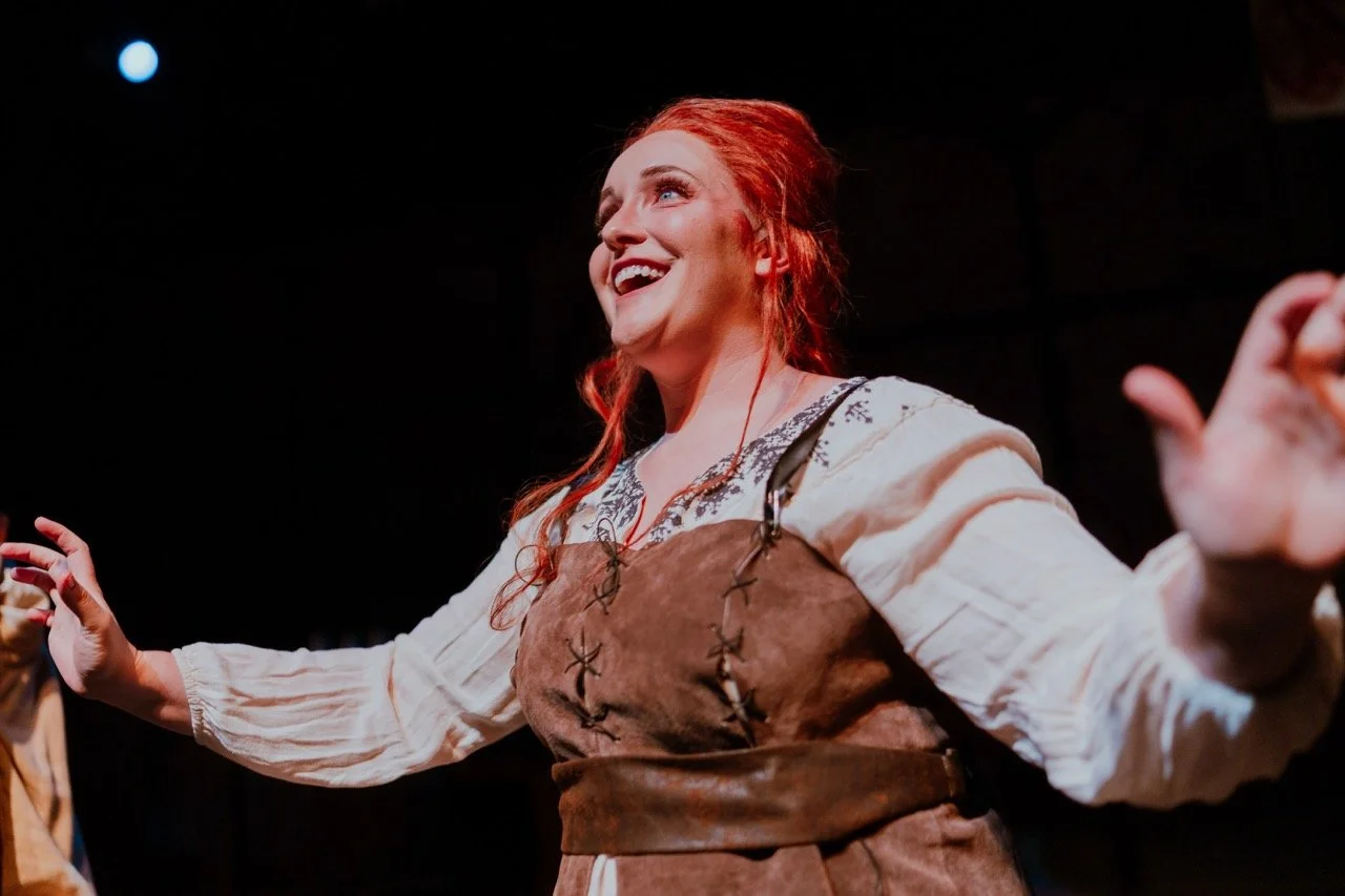 A woman with red hair, wearing a medieval-style costume, performs in a theatrical setting.