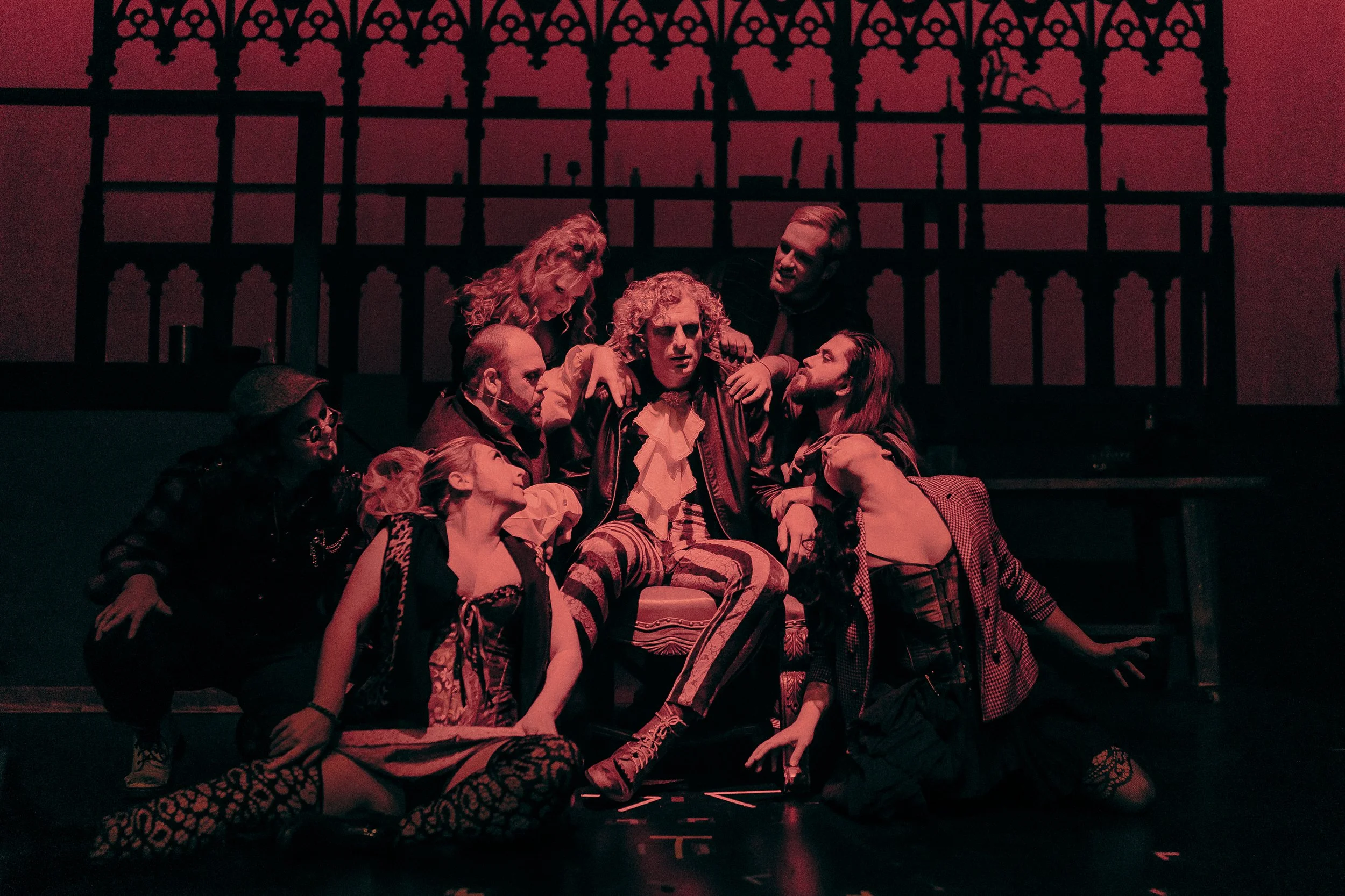 Theatrical performance with a group of actors in dramatic poses under red stage lighting, featuring a central figure dressed in elaborate clothing, seated and surrounded by others.
