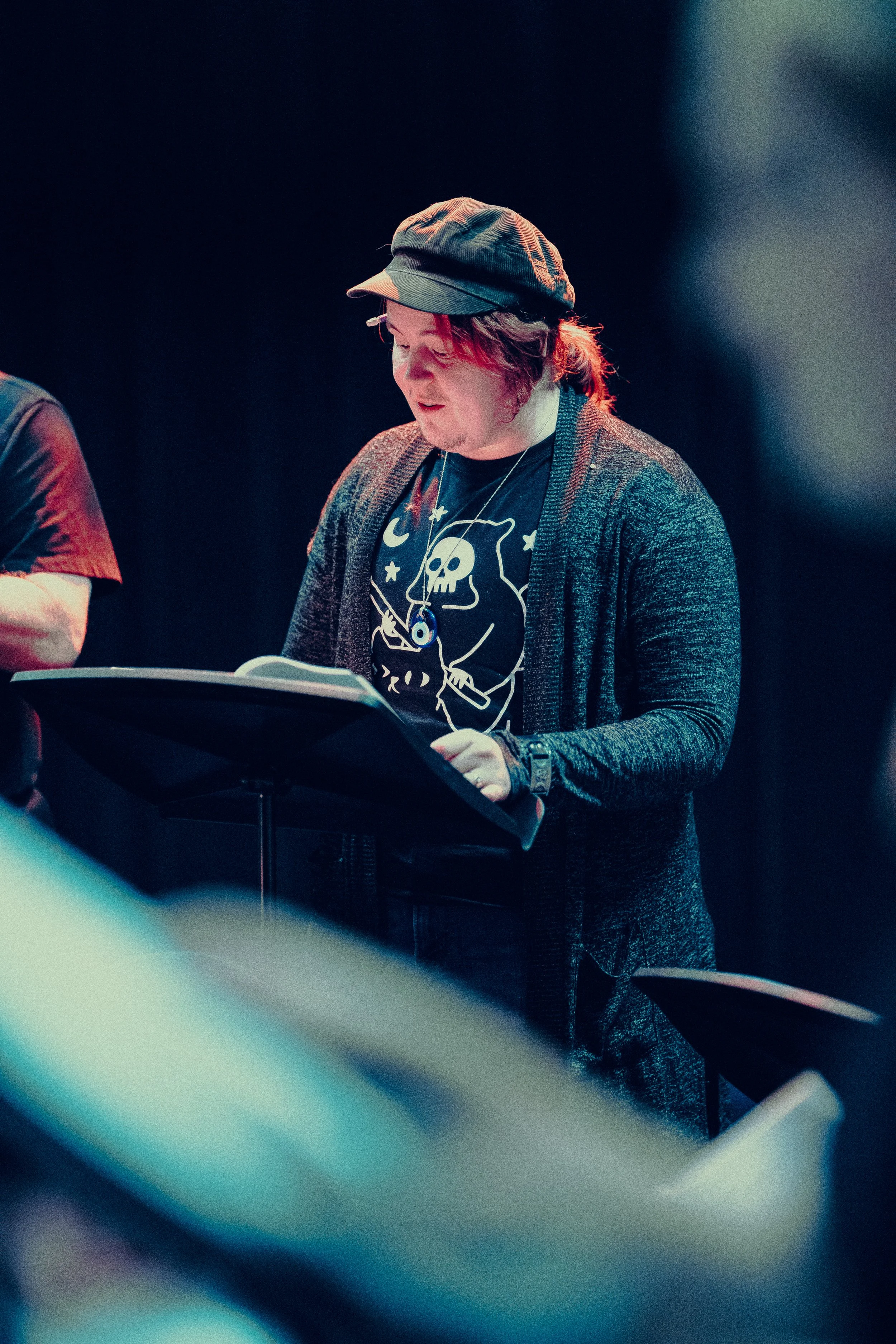 Person wearing a hat and dark sweater, reading script on stand, dimly lit setting.