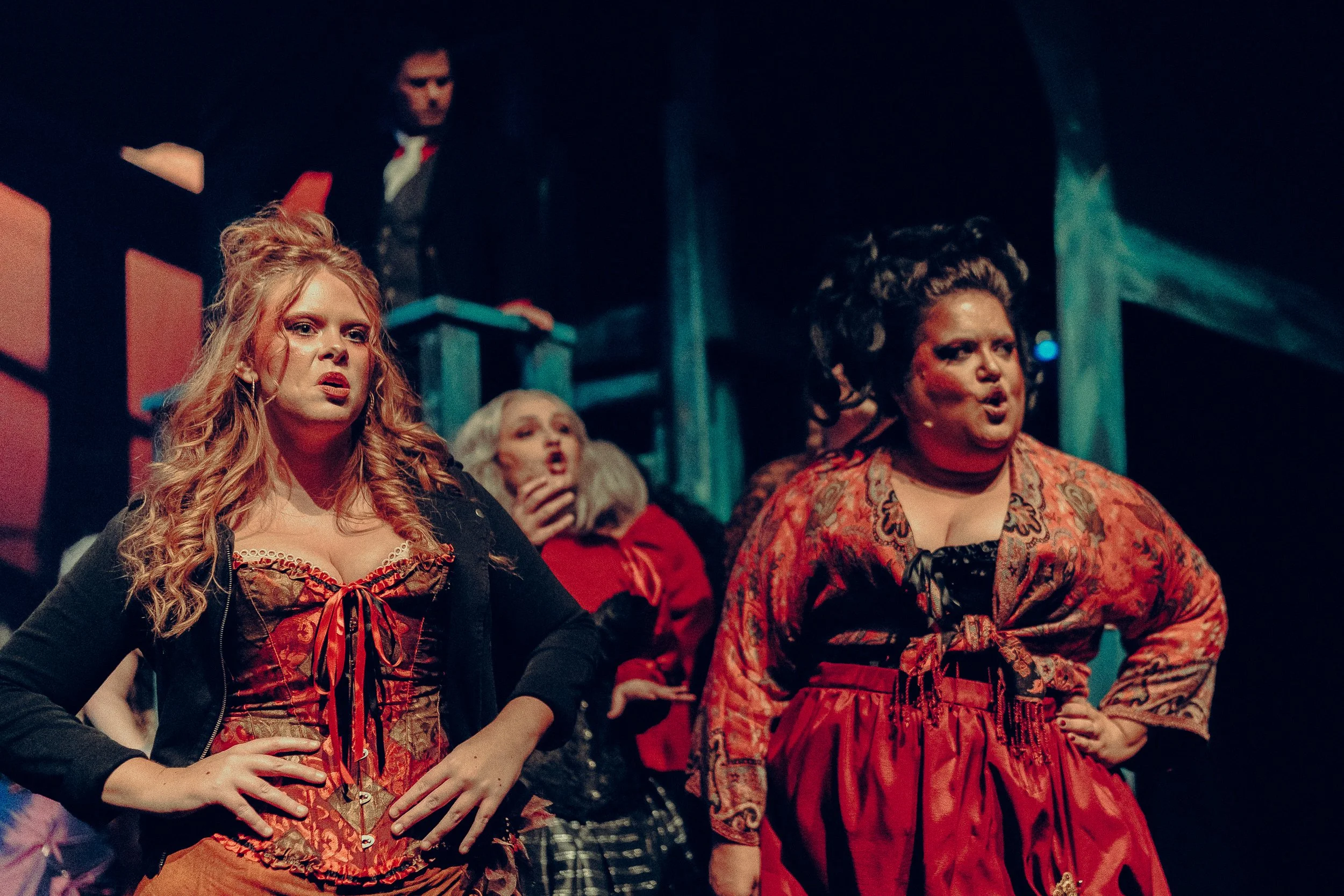 Theater performance with actors in period costumes, dramatic expressions, dim lighting, stage props, red and black attire.