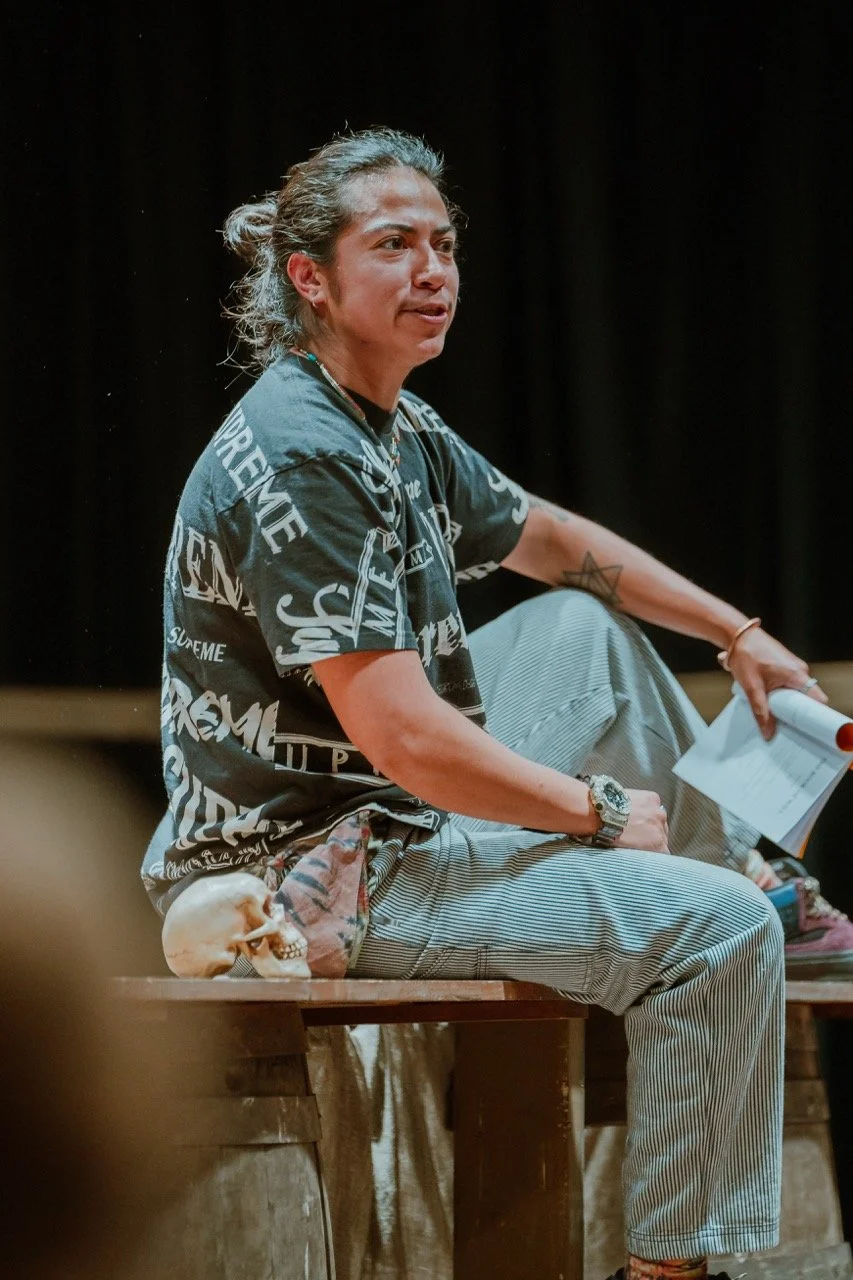 Person sitting on a table, holding papers, wearing a black shirt with text and striped pants, with a skull beside them.