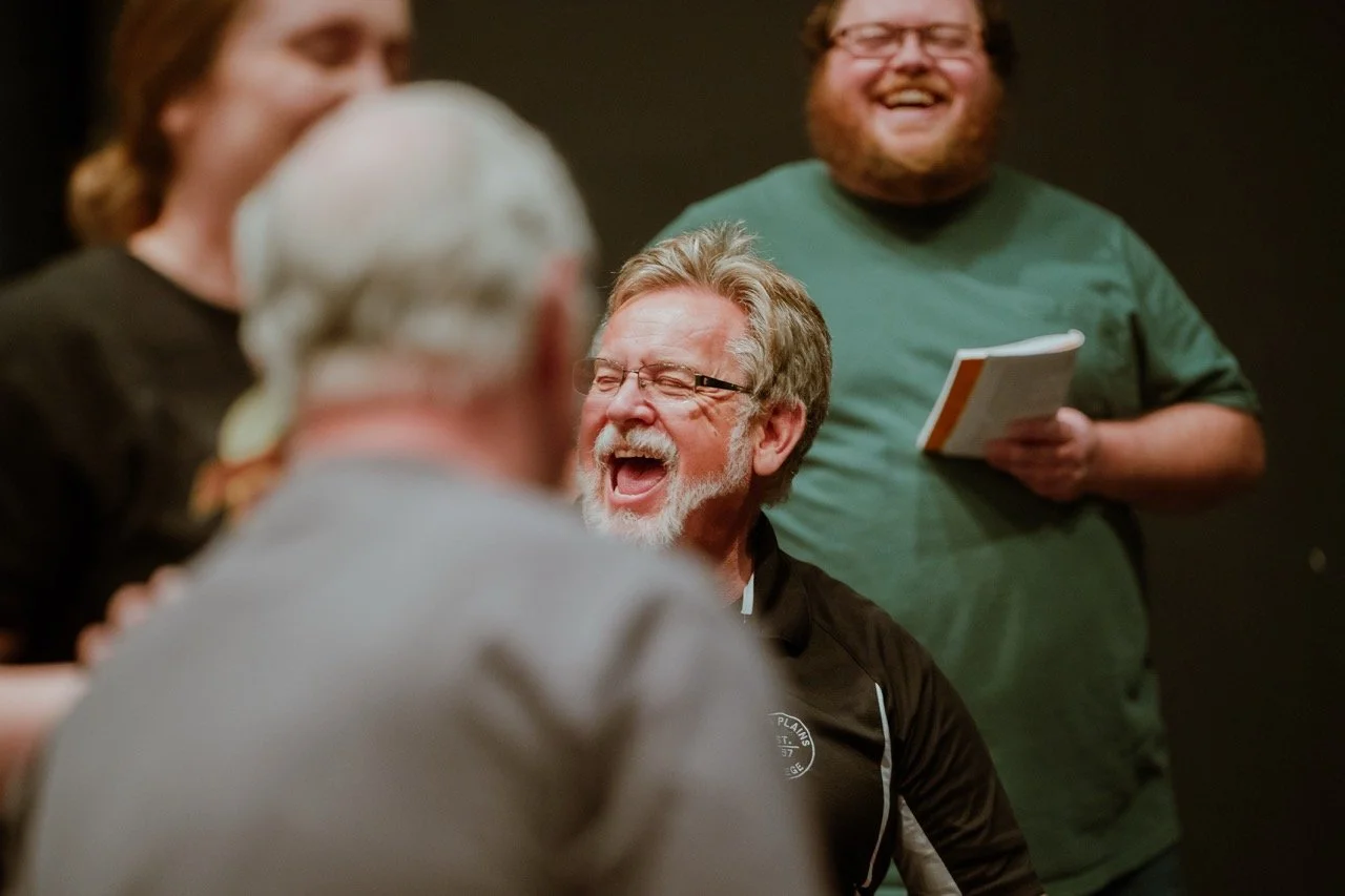 Group of people laughing and holding a book during a gathering or rehearsal in a casual setting.