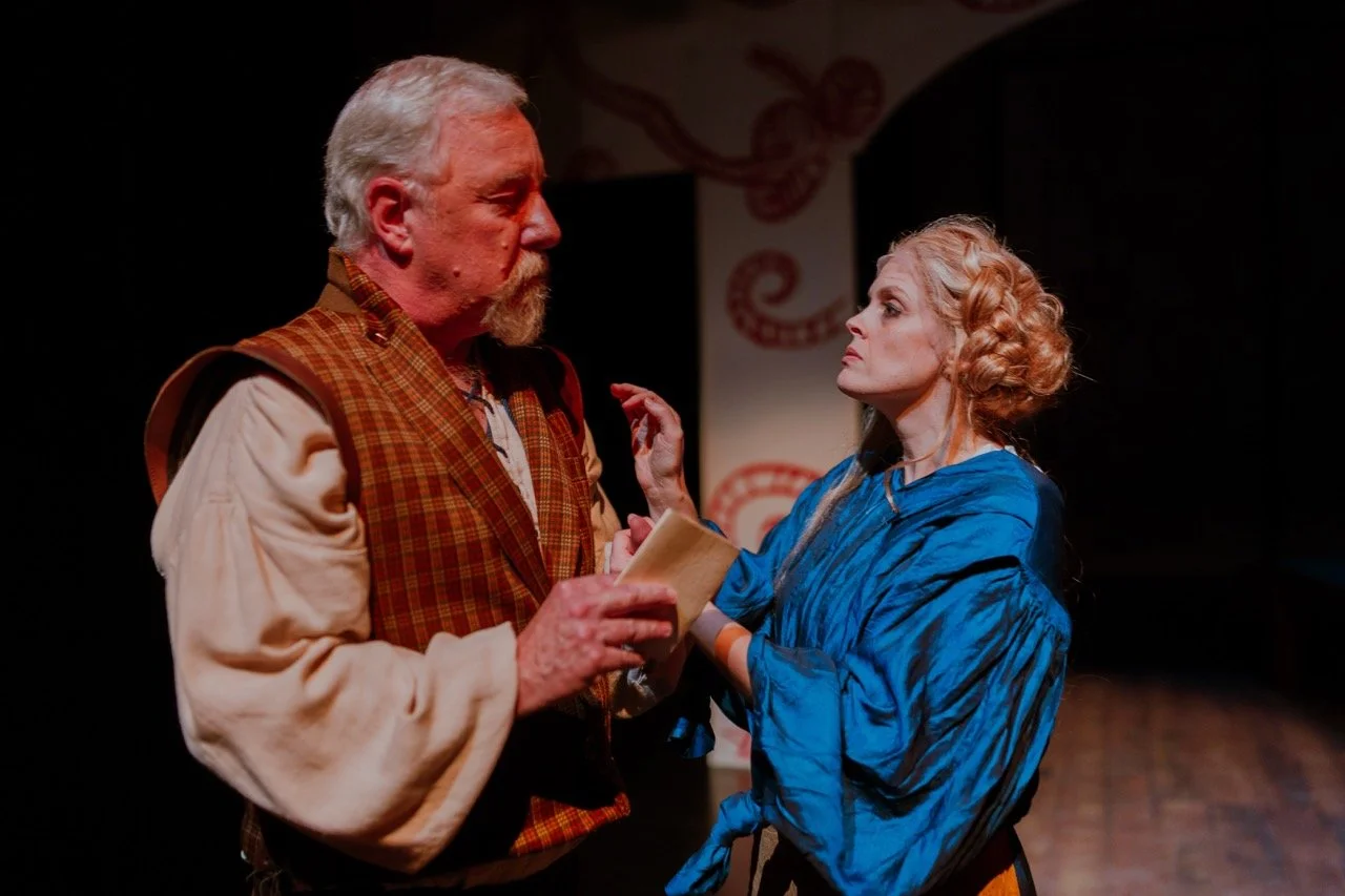 Two actors in period costumes performing a theater scene on stage. One is an older man with white hair in brown, plaid attire, holding a document. The other is a woman with styled blonde hair in a blue dress, looking at him intently.