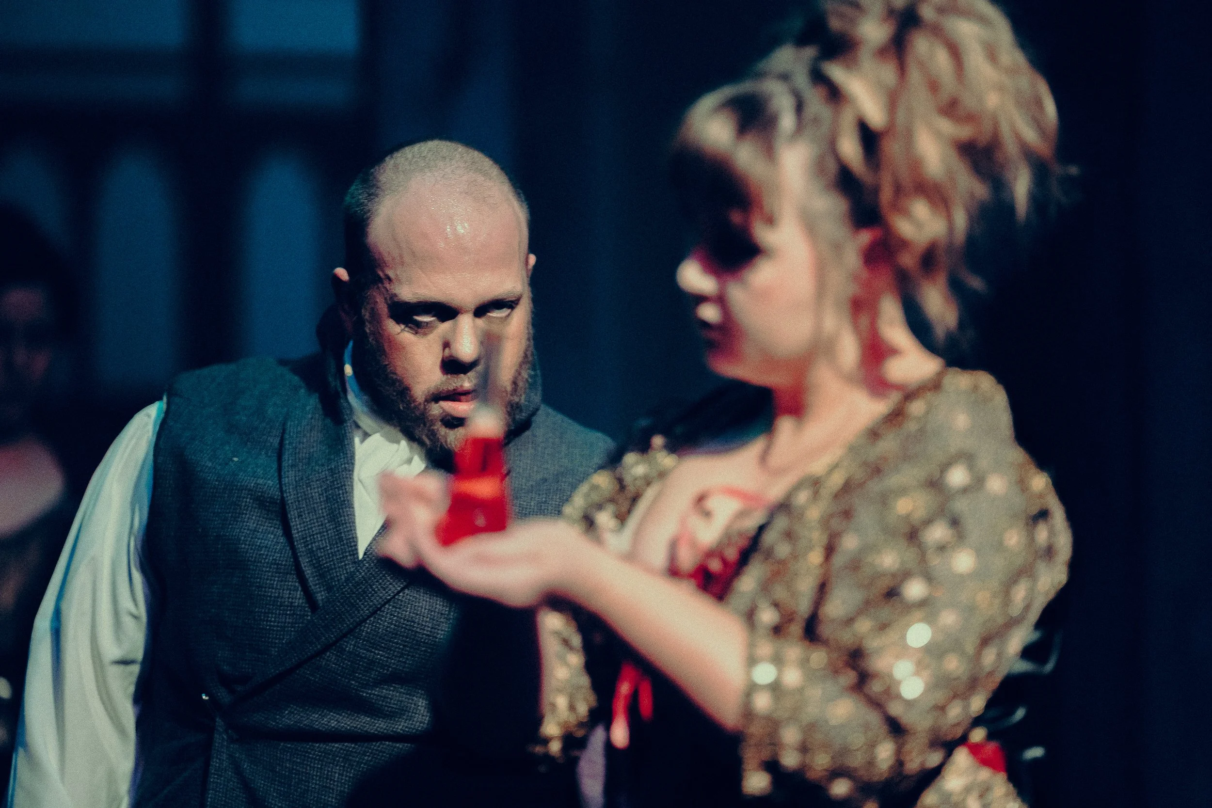 Two actors on stage in dramatic attire, with one man in a suit looking intently at a woman holding a red-colored object, possibly during a theatrical performance.