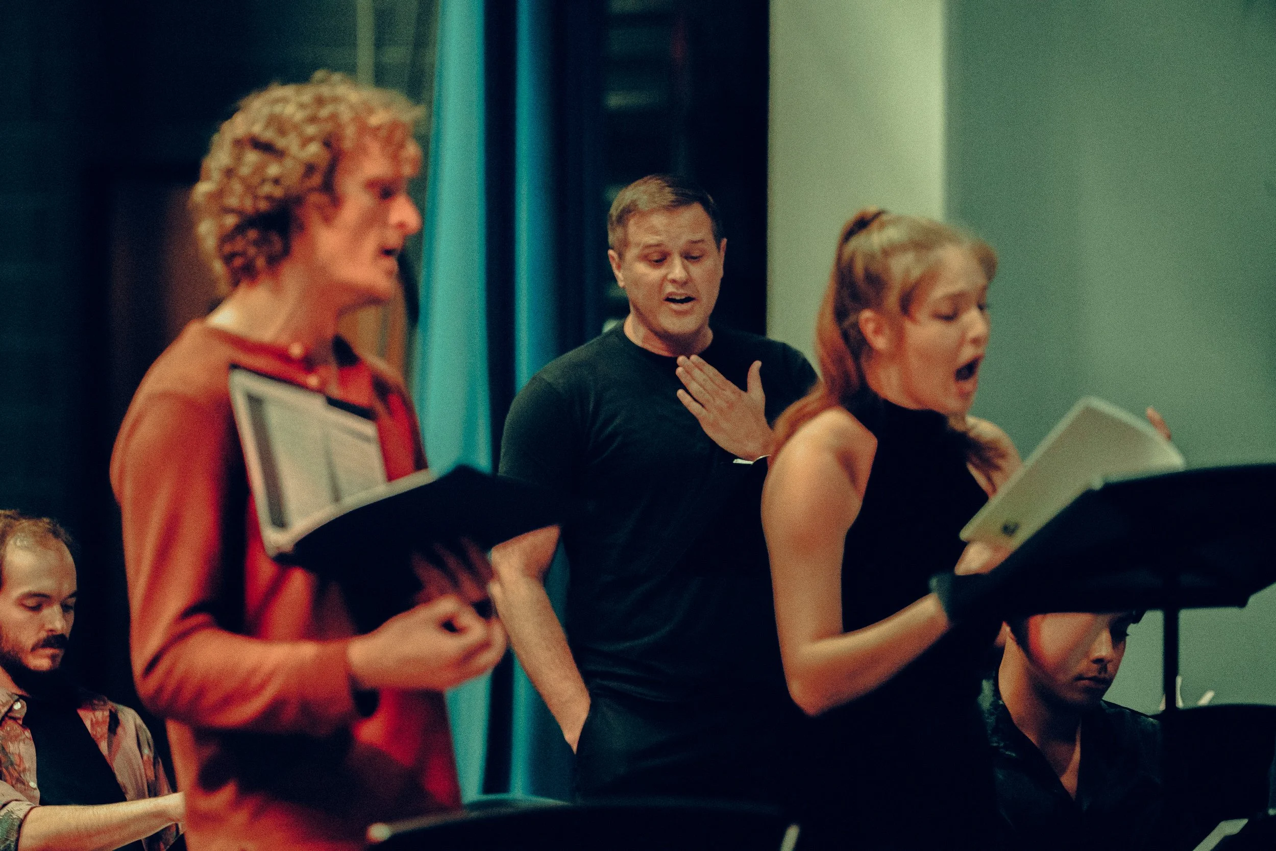 Group of people singing from sheet music, one woman and two men, in a rehearsal setting.
