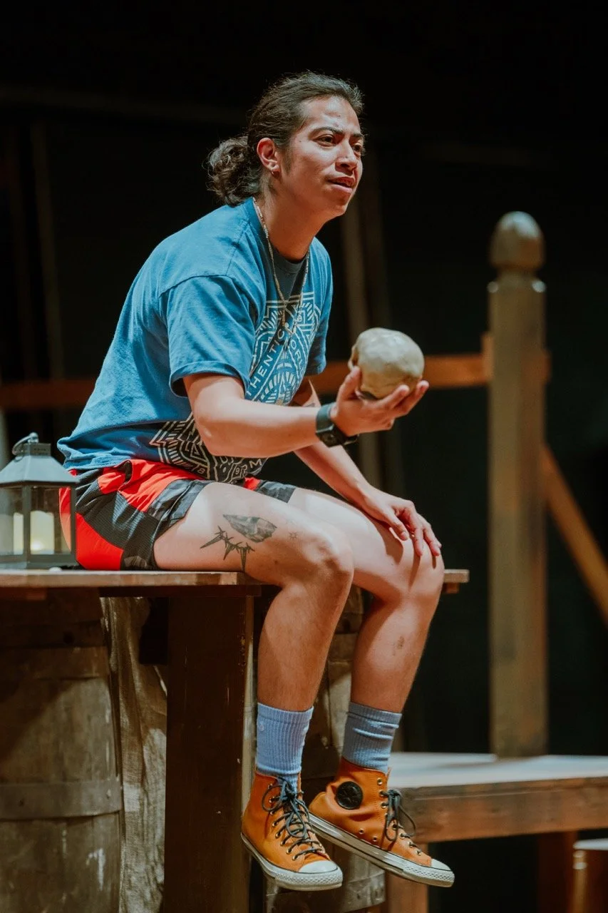 Person sitting on a wooden platform holding a round object, wearing a blue t-shirt, red and black shorts, and yellow high-top sneakers.