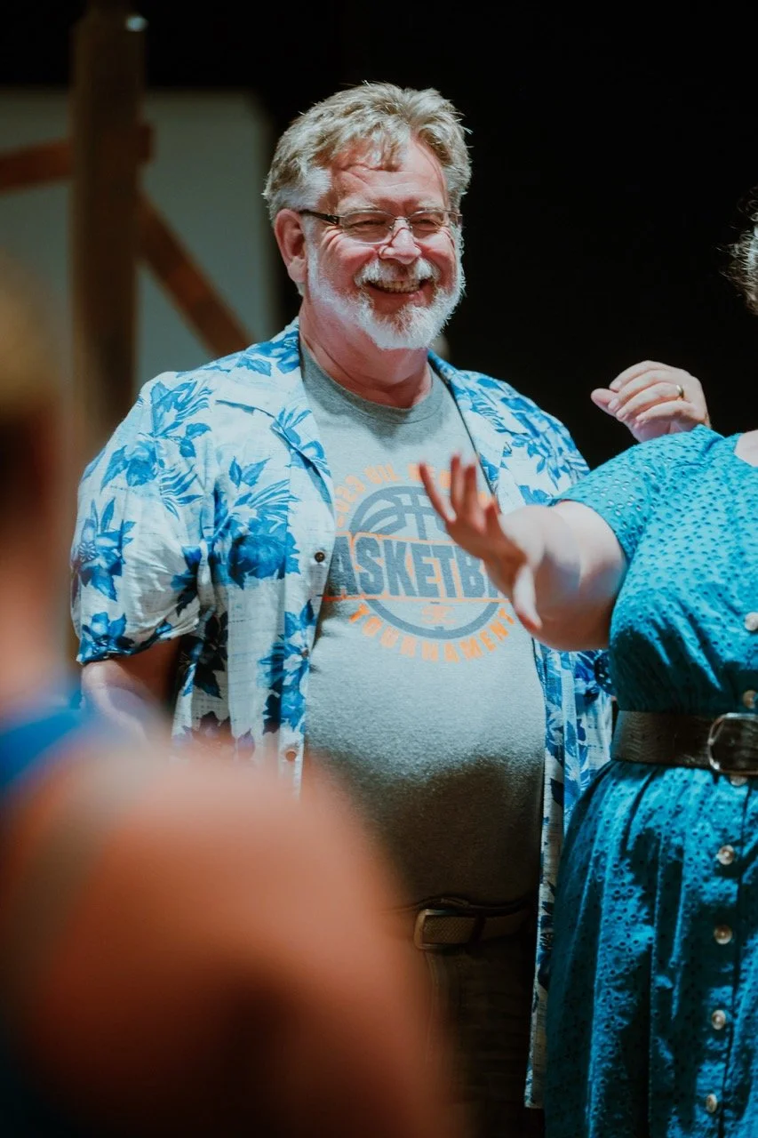 A smiling man with a gray beard wearing glasses, a gray basketball-themed T-shirt, and a blue floral shirt standing indoors with other people partially visible.