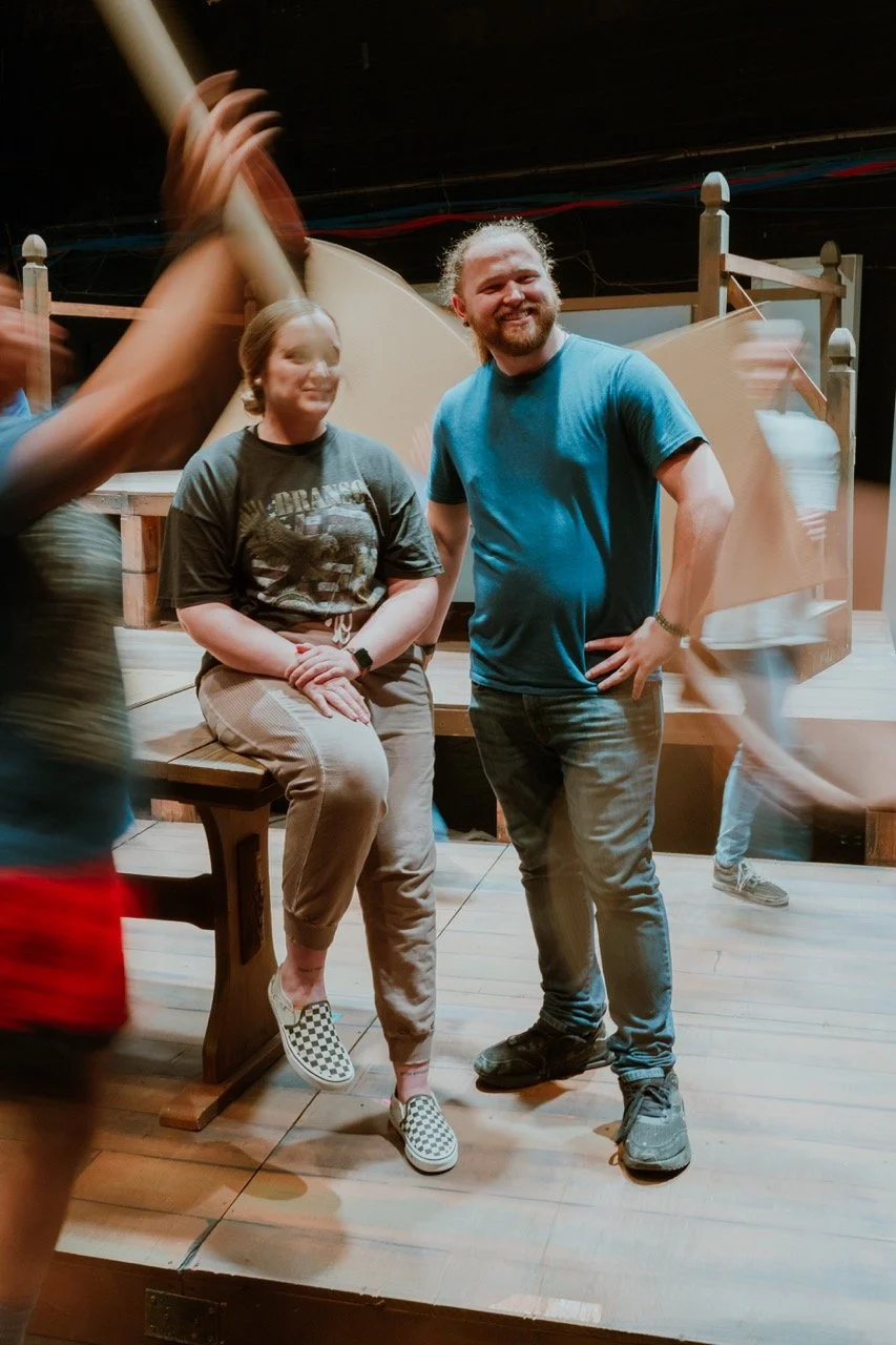 Two people smiling on a stage with motion-blurred individuals around them, wooden stage set in the background.