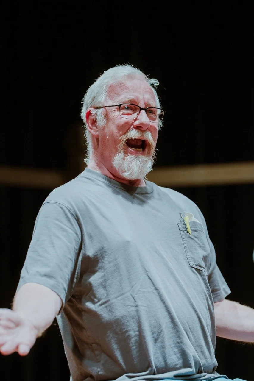 Elderly man with glasses, white beard, and gray shirt speaking passionately on stage.