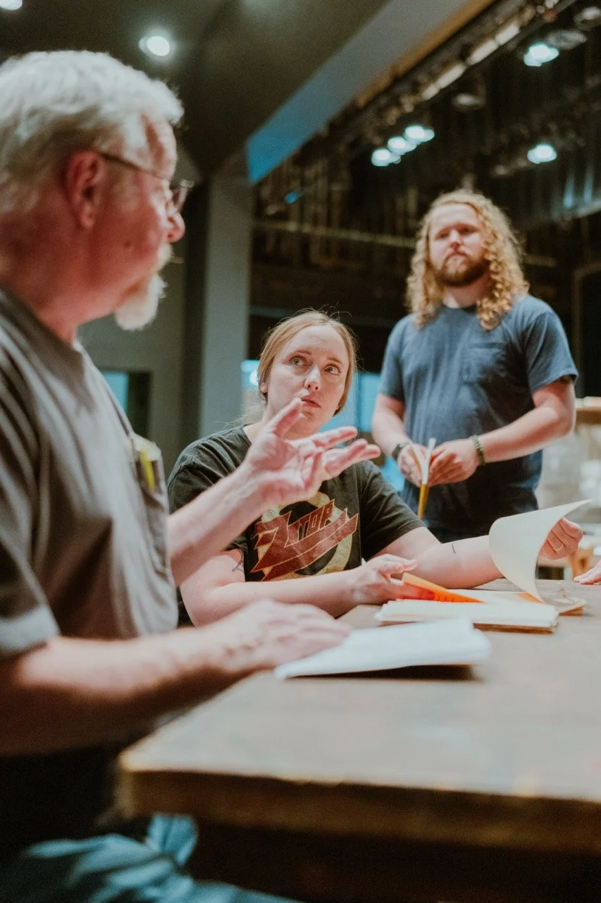 Three people engaged in a discussion or rehearsal, sitting and standing around a wooden table with papers in a dimly lit room.
