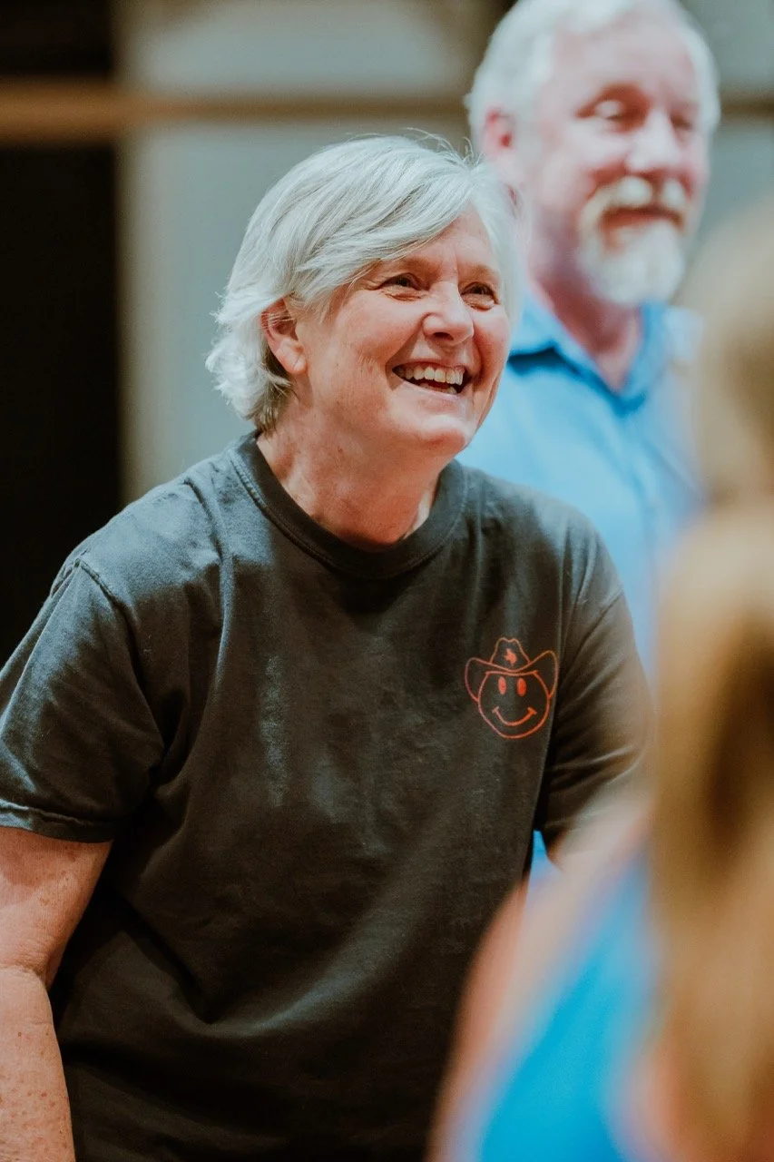 Smiling person with gray hair wearing a T-shirt featuring a smiley face logo, standing indoors with another person blurred in the background.