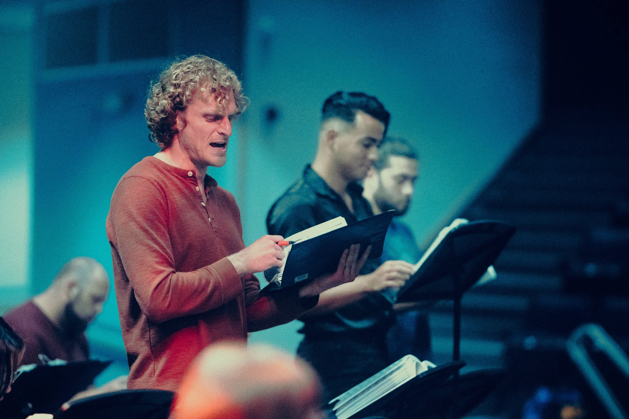 Group of people singing and reading from music sheets in a dimly lit room, possibly during a choir rehearsal.
