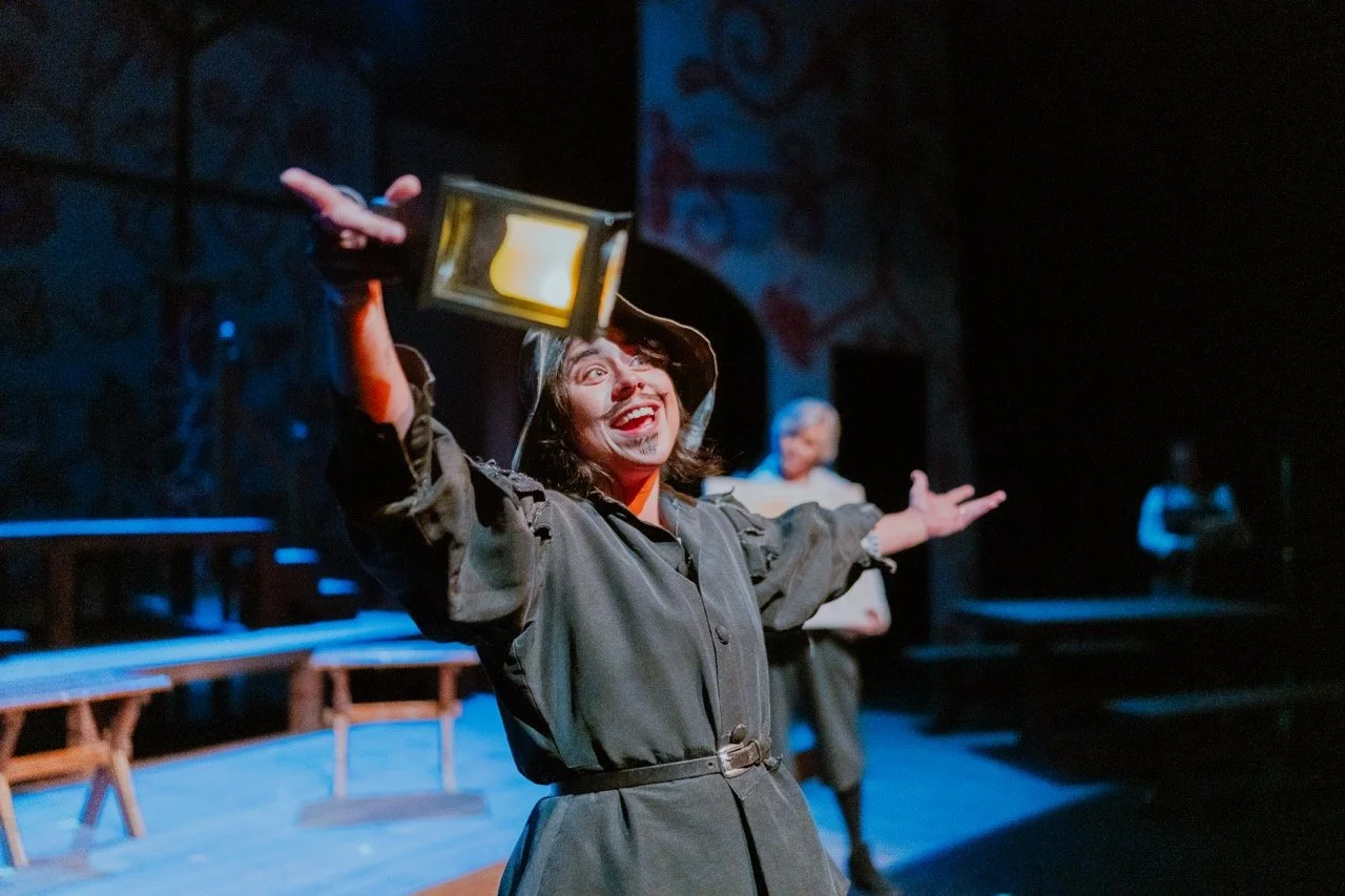 Actor on stage wearing medieval costume, holding a lantern, with an expressive pose and smile, theater setting with wooden benches in background.