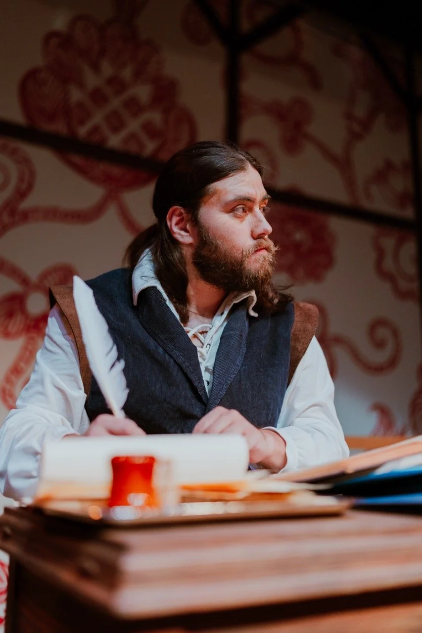 A man with a beard holding a quill pen, sitting at a wooden desk with papers, wearing historical clothing. Background features intricate red patterns.