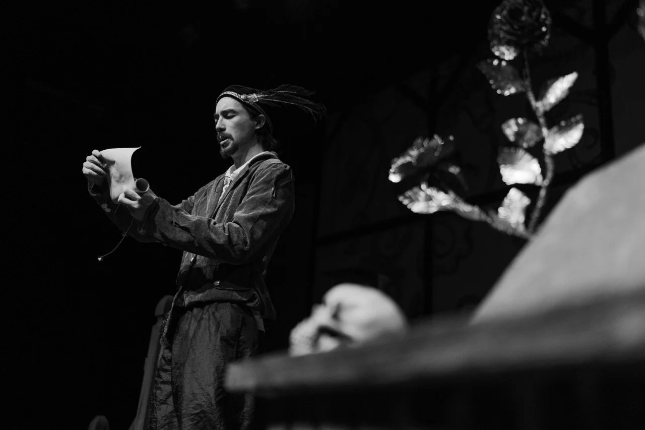 Black and white image of a man in historical costume on stage reading a scroll, with a skull and ornate flower in the foreground.