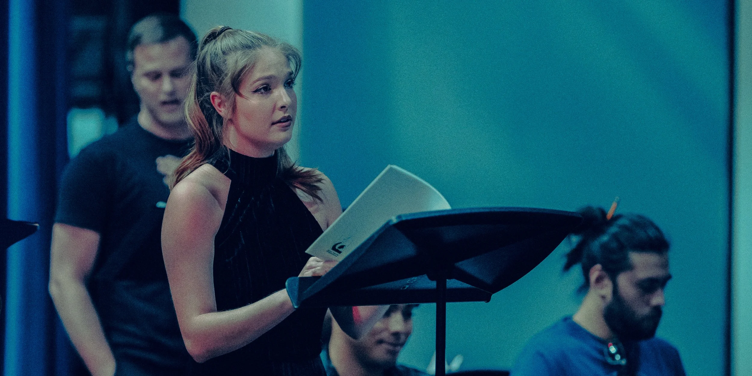 A woman singing with a sheet music stand, with two men in the background.