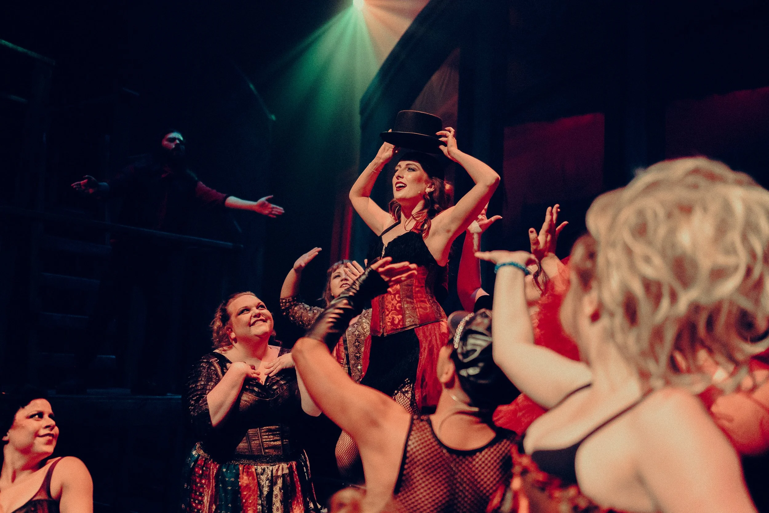 Performers on stage in theatrical costumes, with one person wearing a top hat and red corset surrounded by others gesturing energetically.