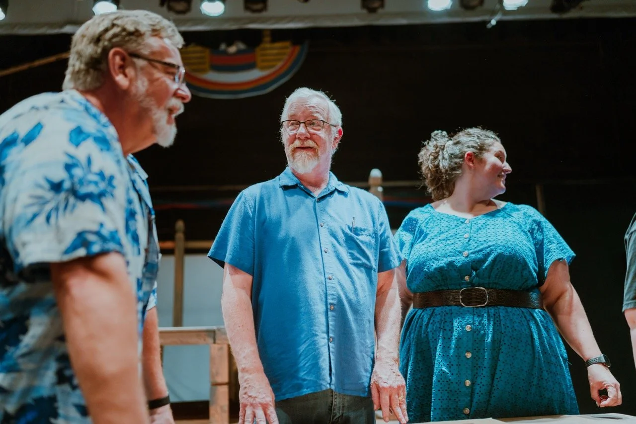 Three people wearing blue clothing are standing and talking in a room with stage lights overhead.
