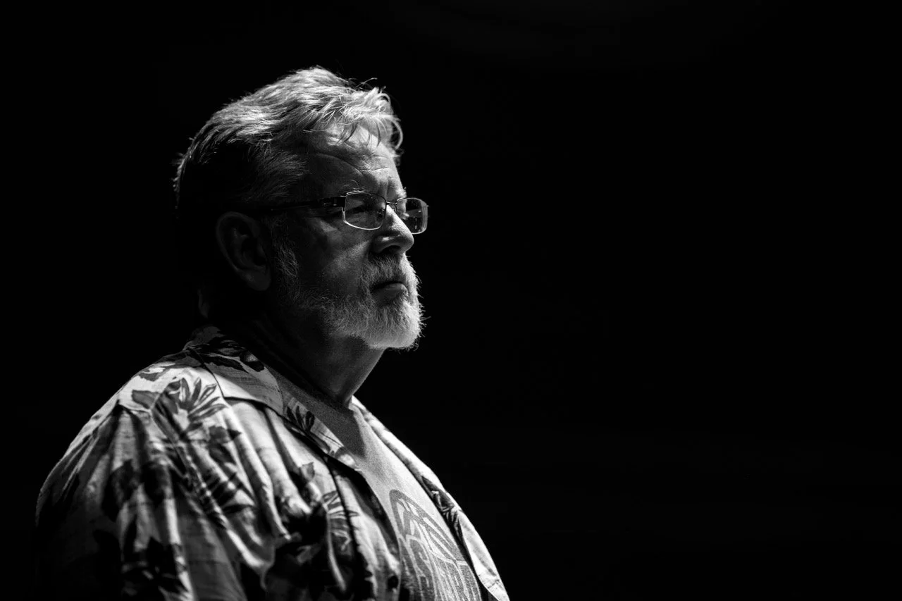 Black and white portrait of a bearded person wearing glasses and a patterned shirt, looking to the side against a dark background.