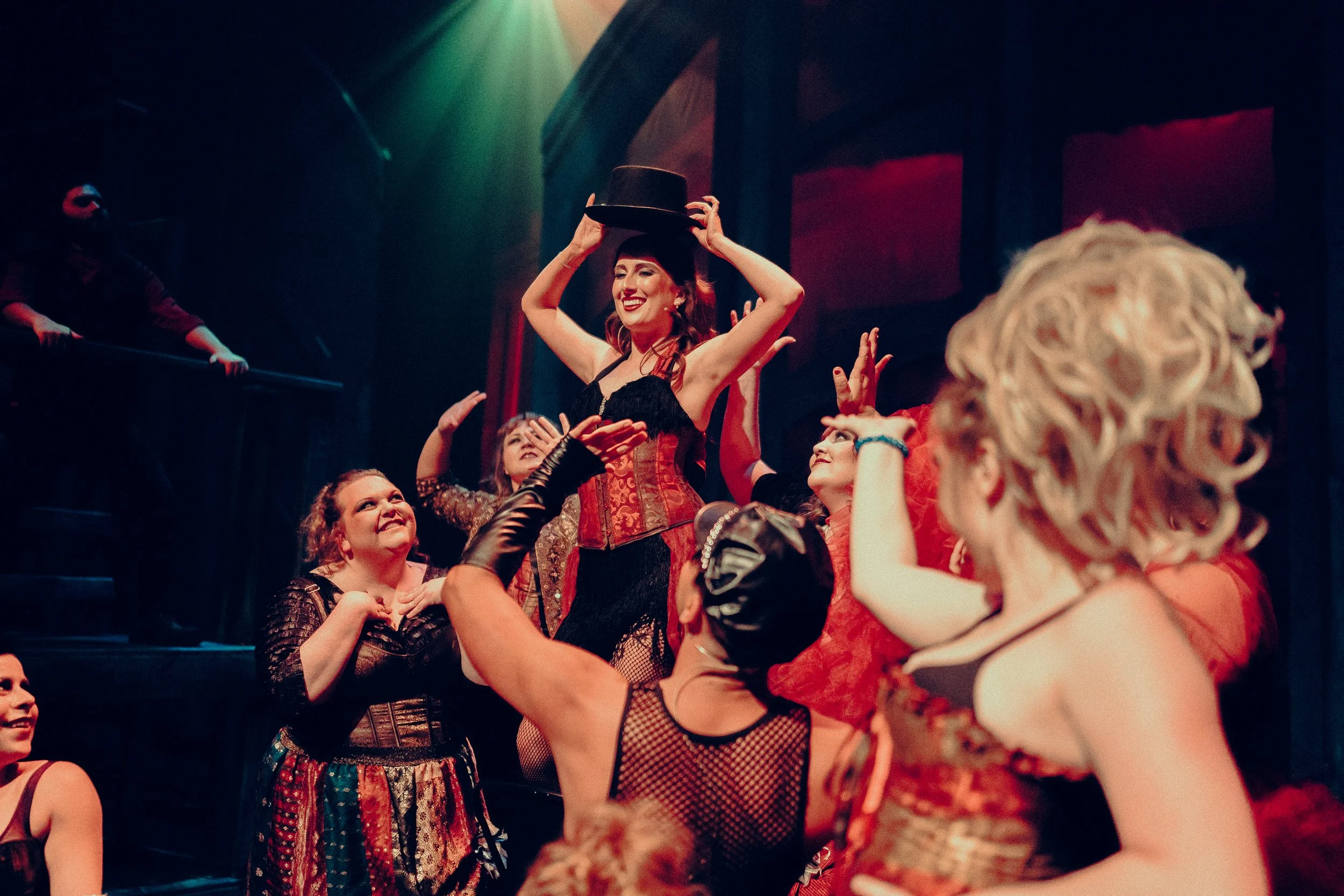 A scene from a theatrical performance featuring performers in Victorian-style costumes with corsets and top hats. One performer stands out, holding a top hat above her head, surrounded by others cheering and gesturing in a stage setting with dramatic