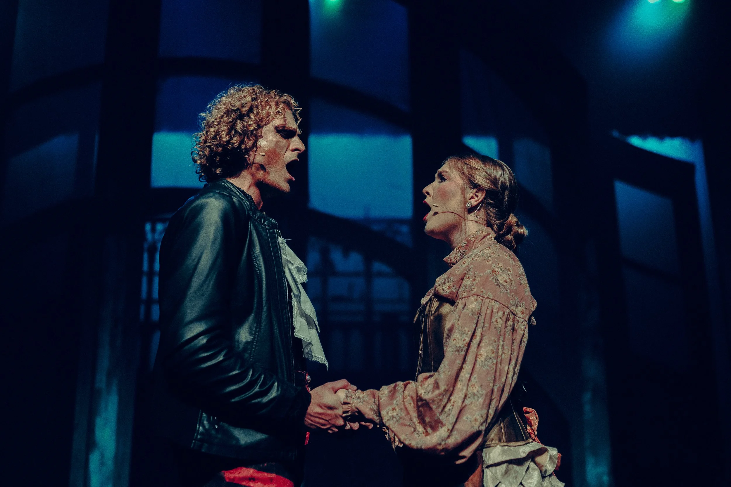 A man and a woman performing on stage in period costumes, holding hands and singing passionately. The scene is dimly lit with a blue background, suggesting a dramatic theater production.