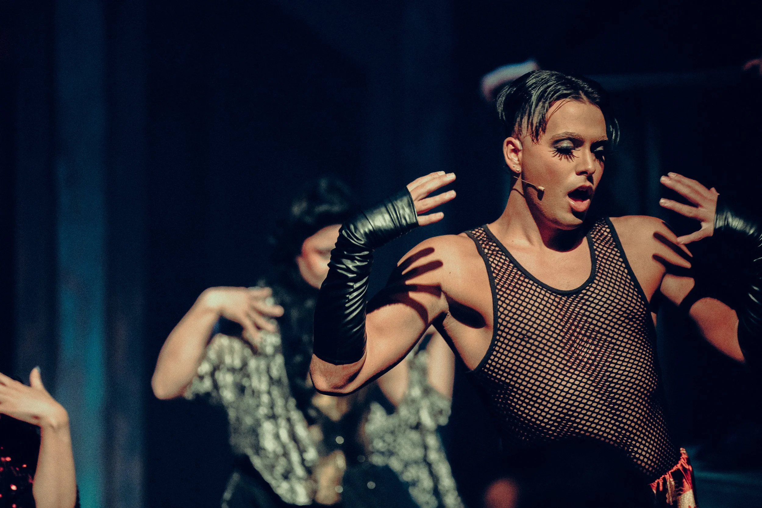 Performer on stage wearing a fishnet top and black gloves, singing or acting with expressive gestures at a theatrical event.