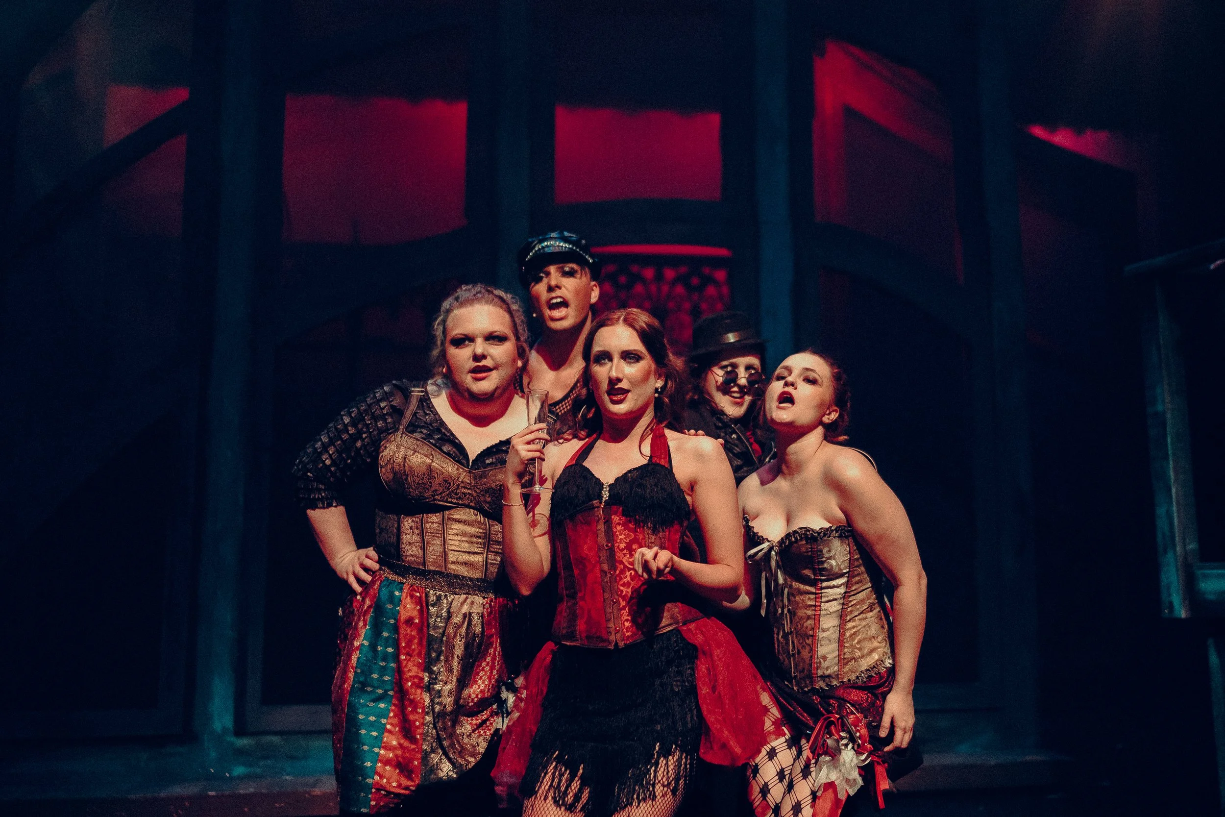 Five people in theatrical costumes, including corsets and lace, standing on stage with a dimly lit, red and blue background.