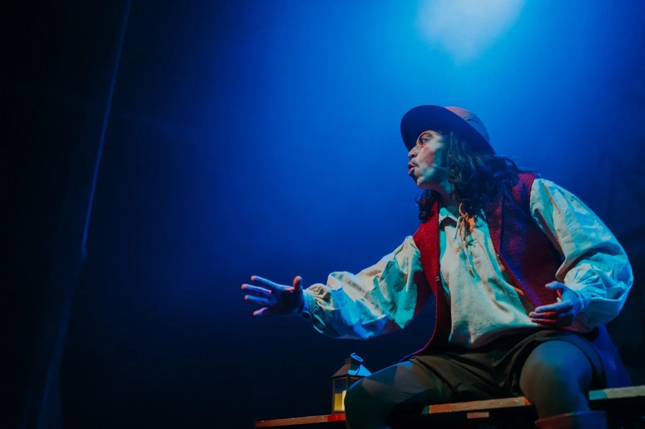 Performer on stage under blue spotlight, wearing a red vest and hat, gesturing expressively.