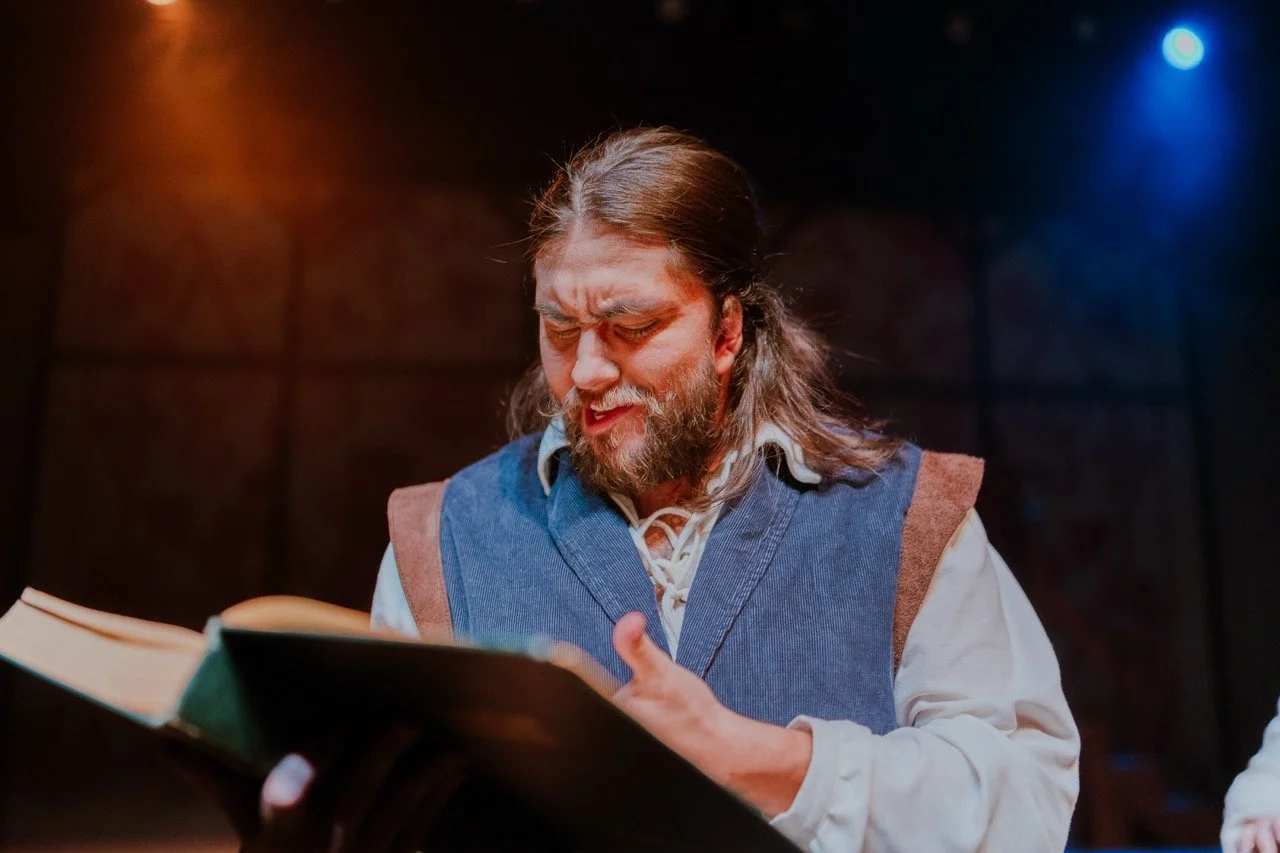 A man with long hair and a beard reading from a book on stage under dramatic lighting.