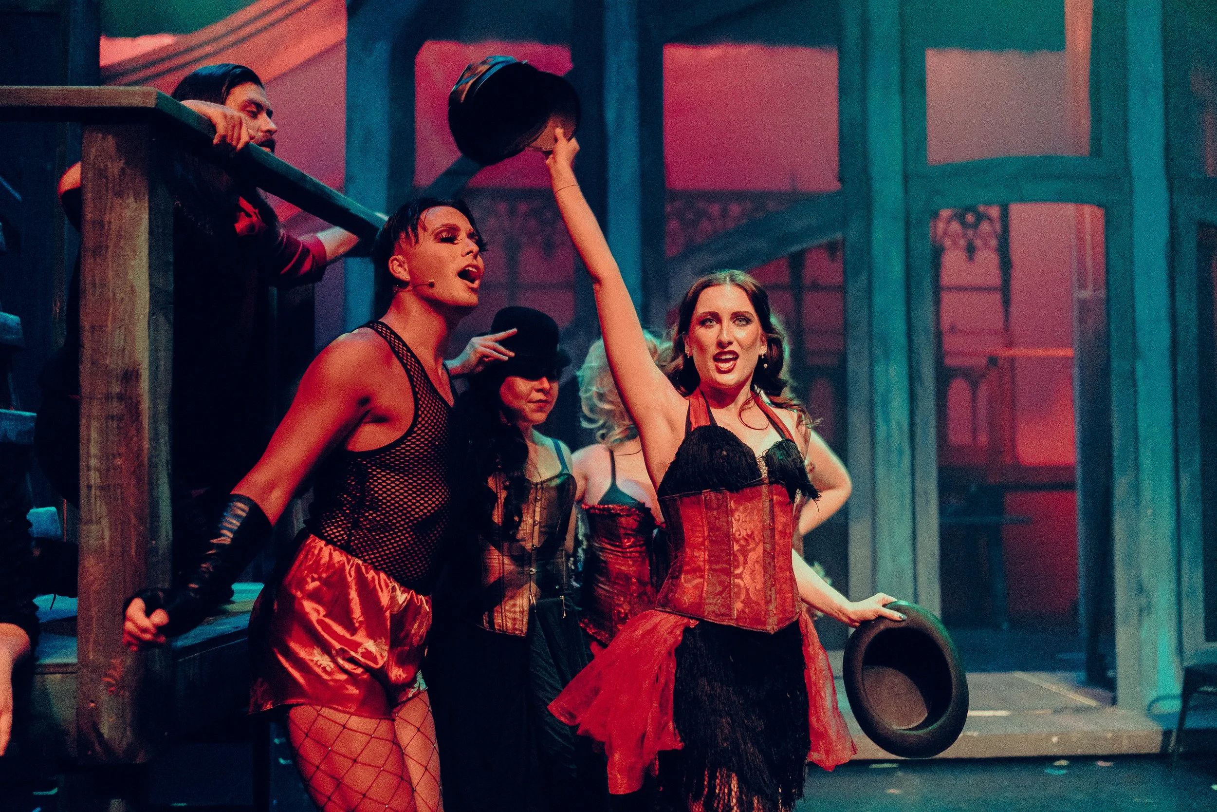 A theatrical performance with actors in vibrant costumes, holding hats, set on a stage with dramatic lighting.