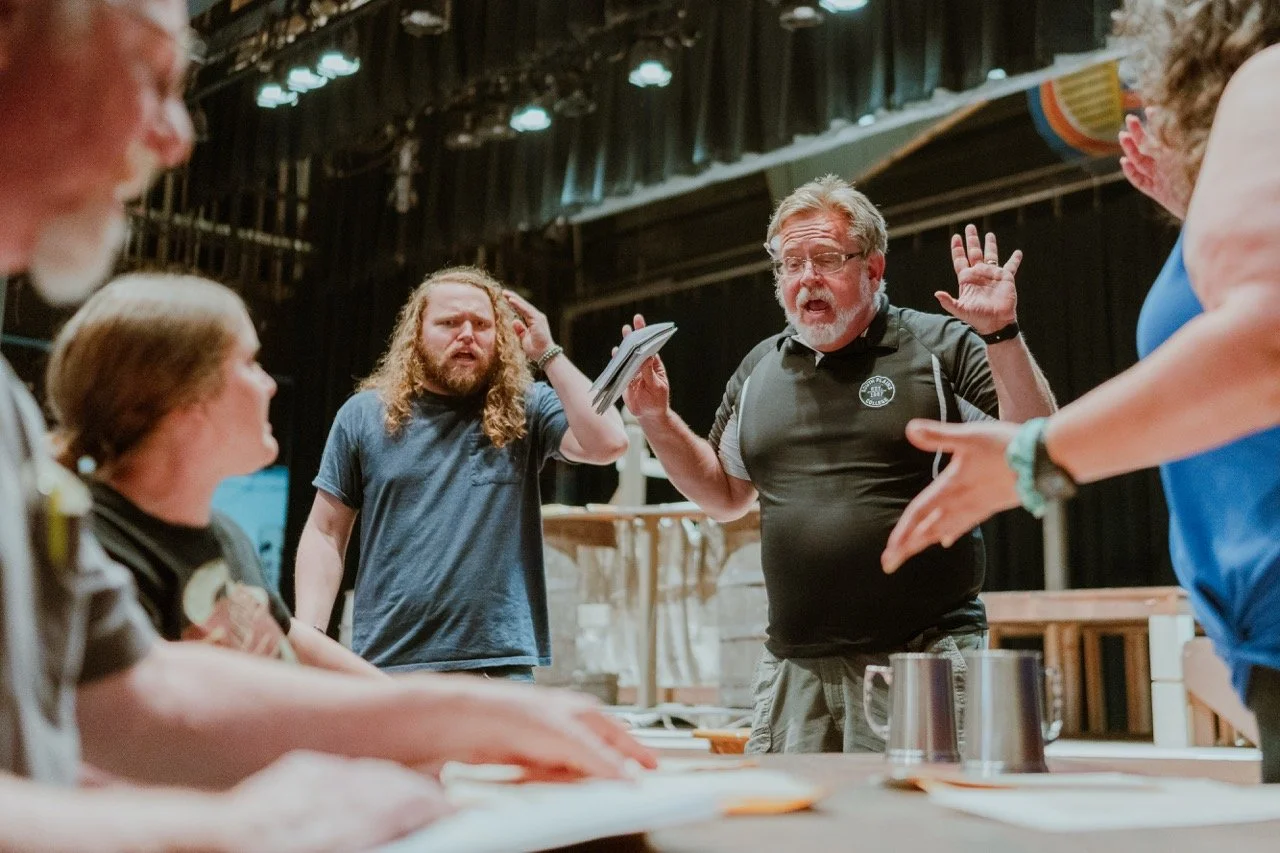 Group of people involved in a theater rehearsal, with one man holding a script and speaking animatedly, while others listen and react on stage.