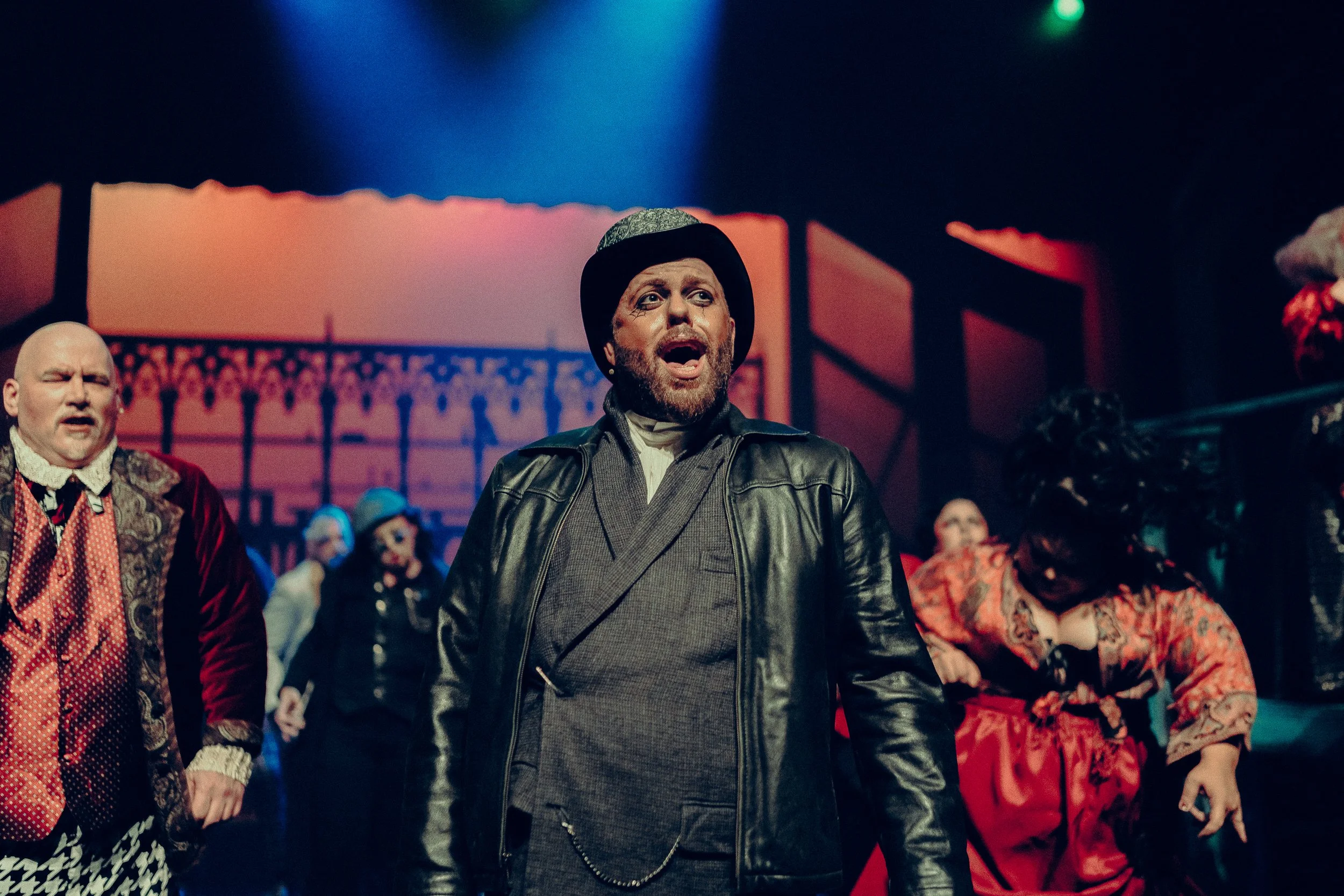 Theatrical performance featuring a man in a leather jacket and bowler hat singing on stage, with other performers wearing period costumes in the background against a dramatic set.
