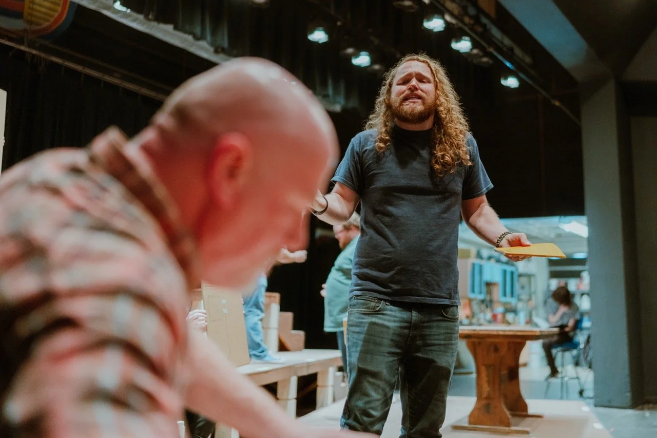 Two people in a theater setting, one with long hair holding a paper, and another blurred in the foreground.